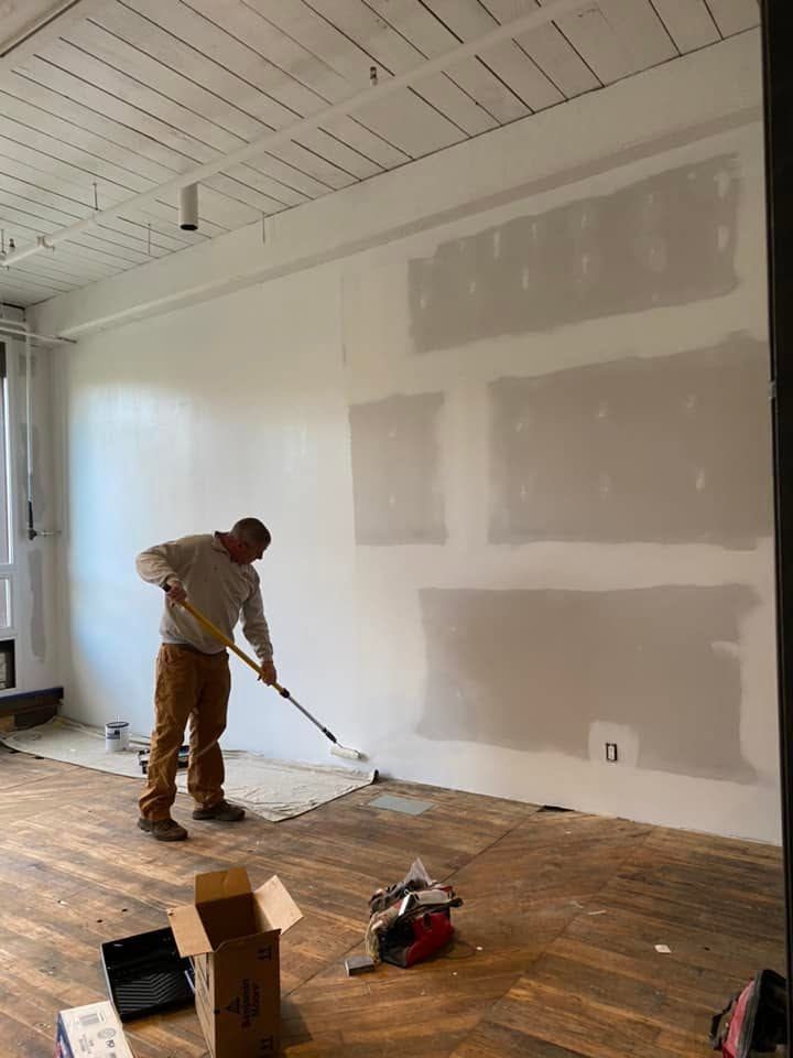 Person painting a white interior wall in a room with patchy drywall and wood floor.