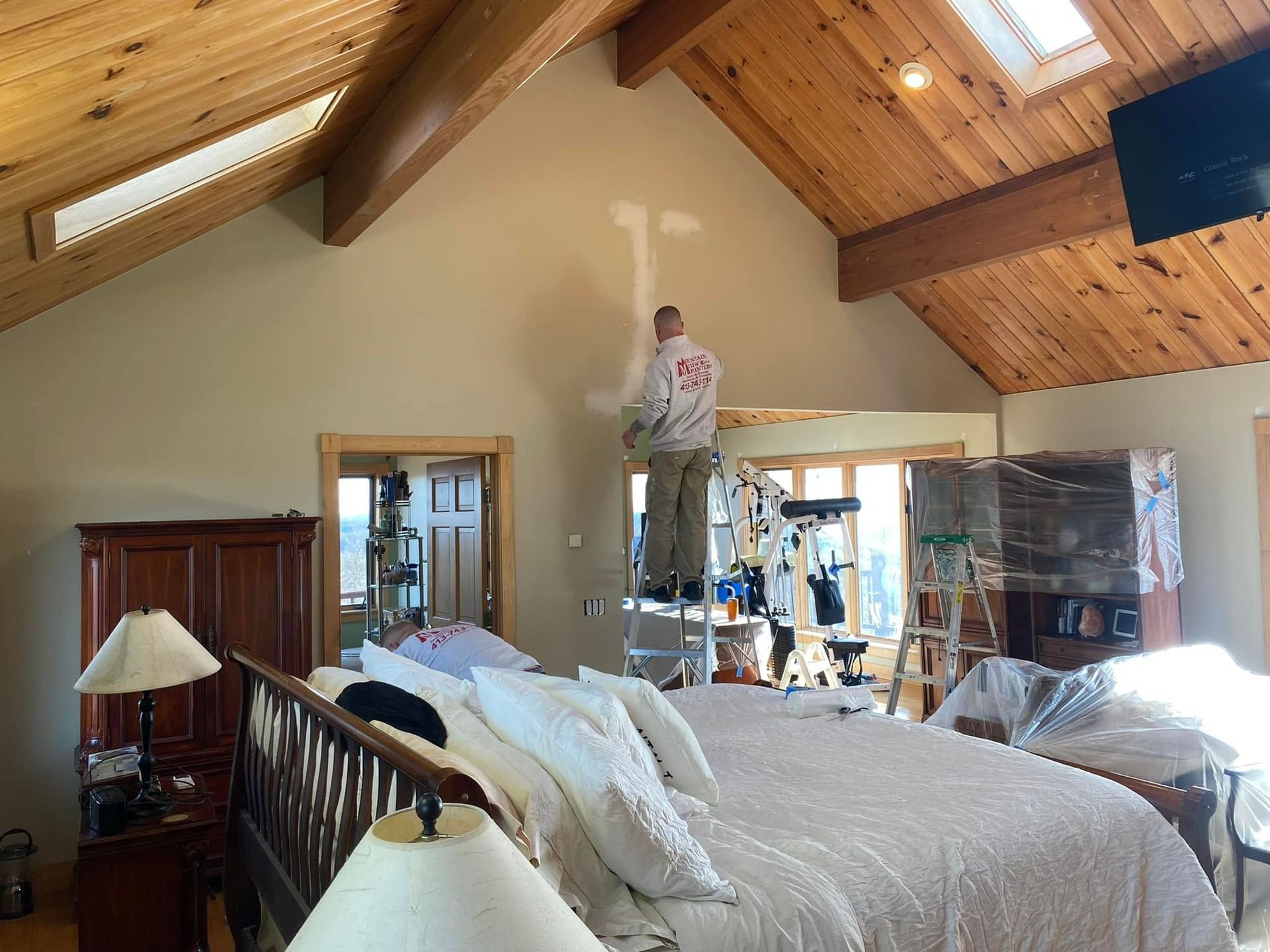 Man standing on a ladder in a bedroom with vaulted wood ceiling during renovation, furniture covered in plastic
