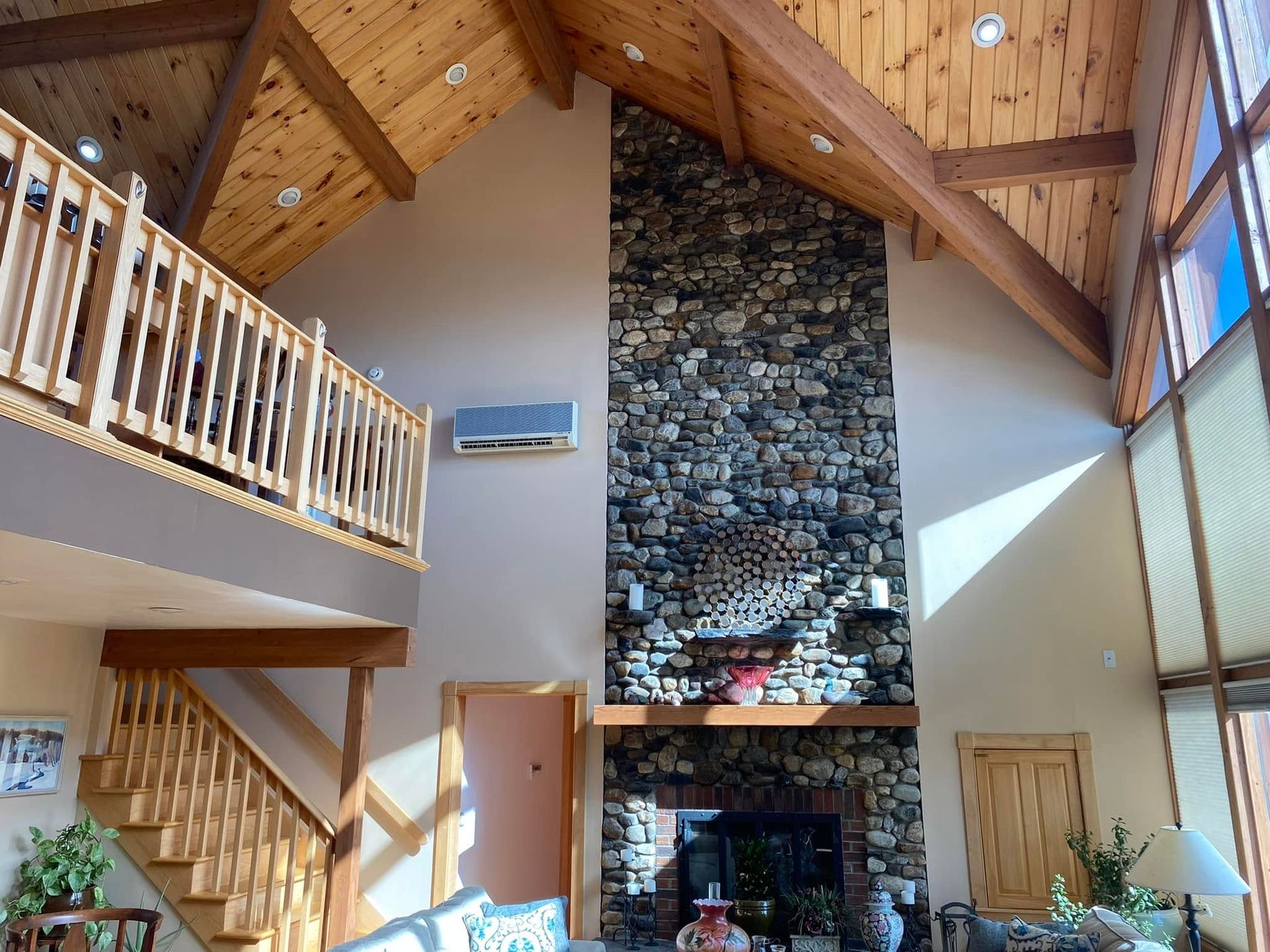 Cozy living room with vaulted wood ceiling, stone fireplace, staircase, and large windows