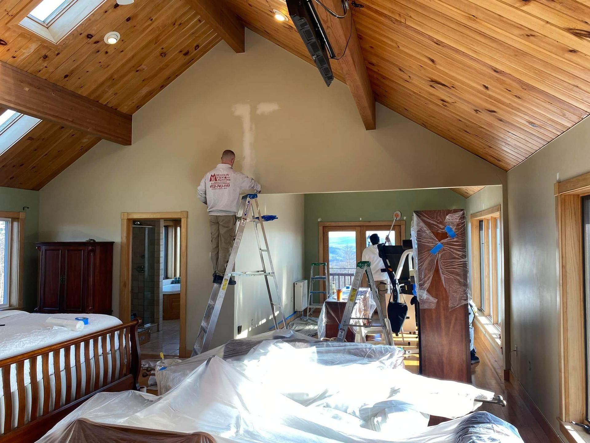 Workers on ladders painting a vaulted bedroom with a wood ceiling and plastic-covered floor.