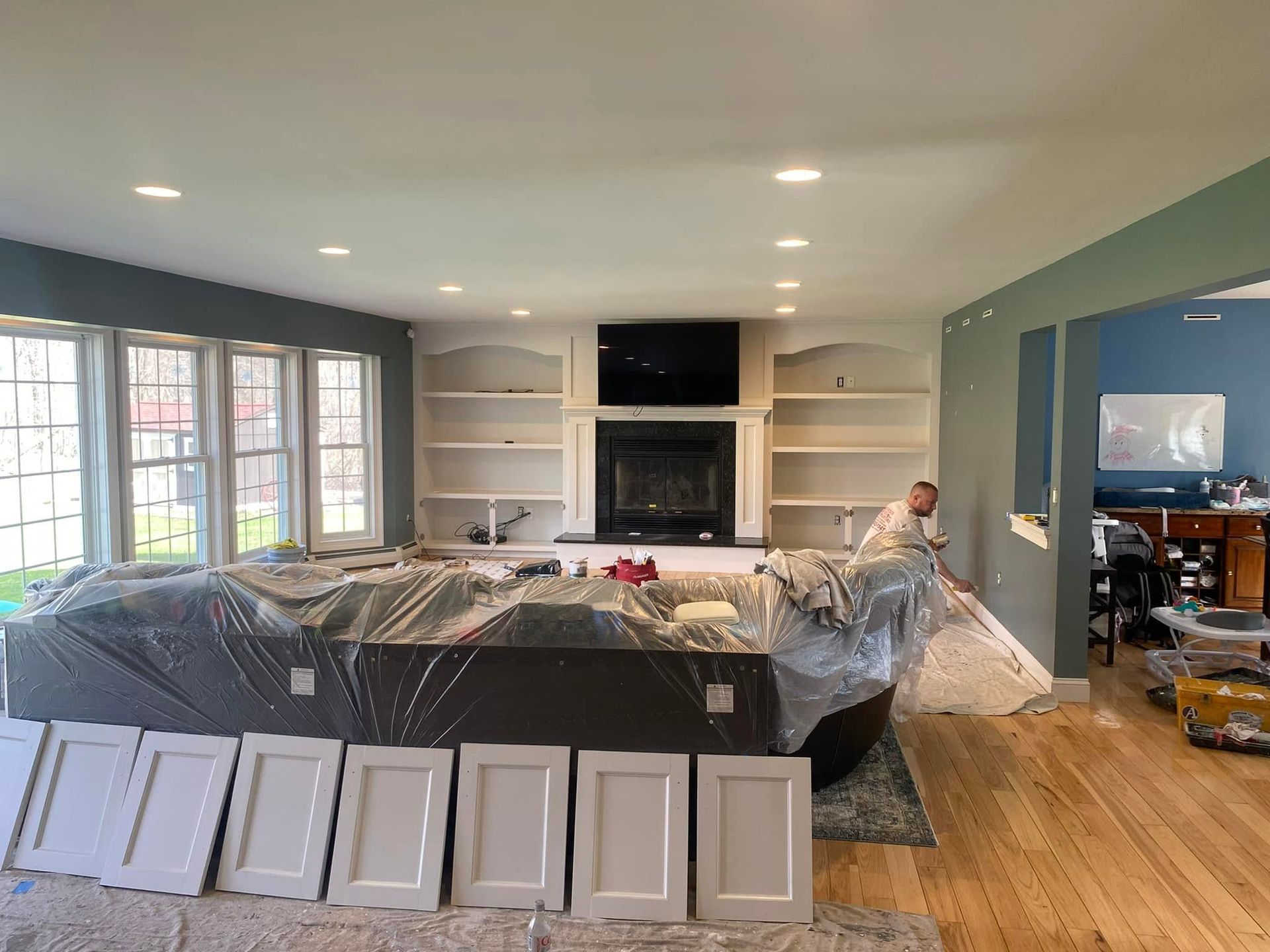 Kitchen under renovation with cabinets, plastic-covered island, and a worker installing trim.