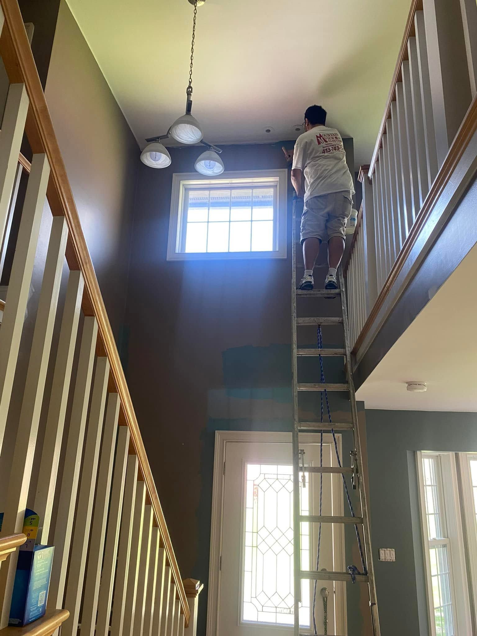 Person standing on a ladder in a two-story hallway, reaching toward the ceiling near a window.