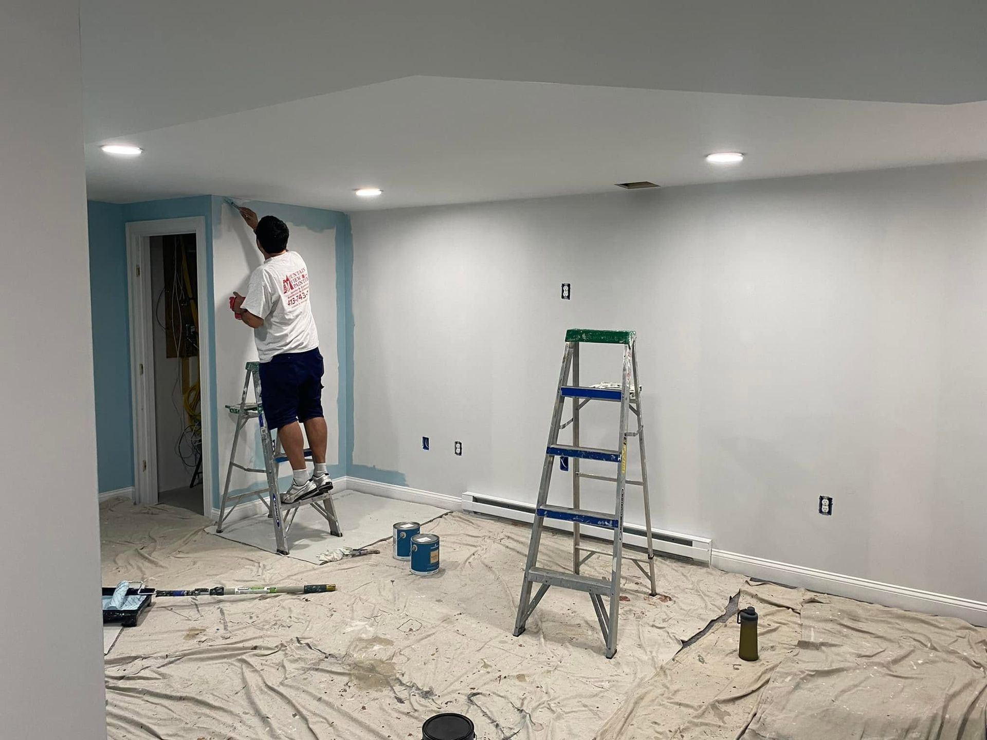 Two workers painting a white room with ladders and drop cloths on the floor.