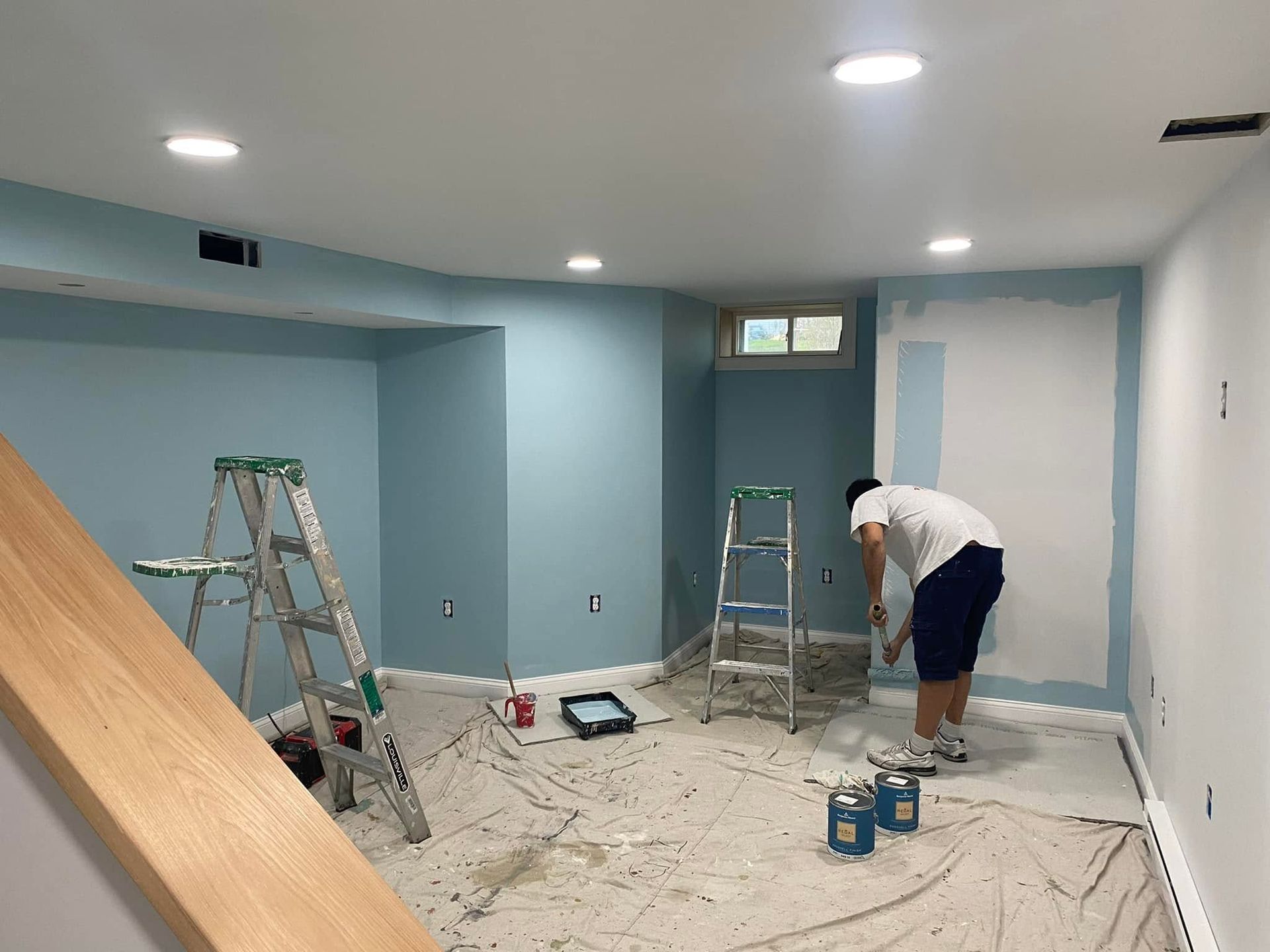 Person painting a basement room light blue, with ladders, tools, and drop cloths on the floor.