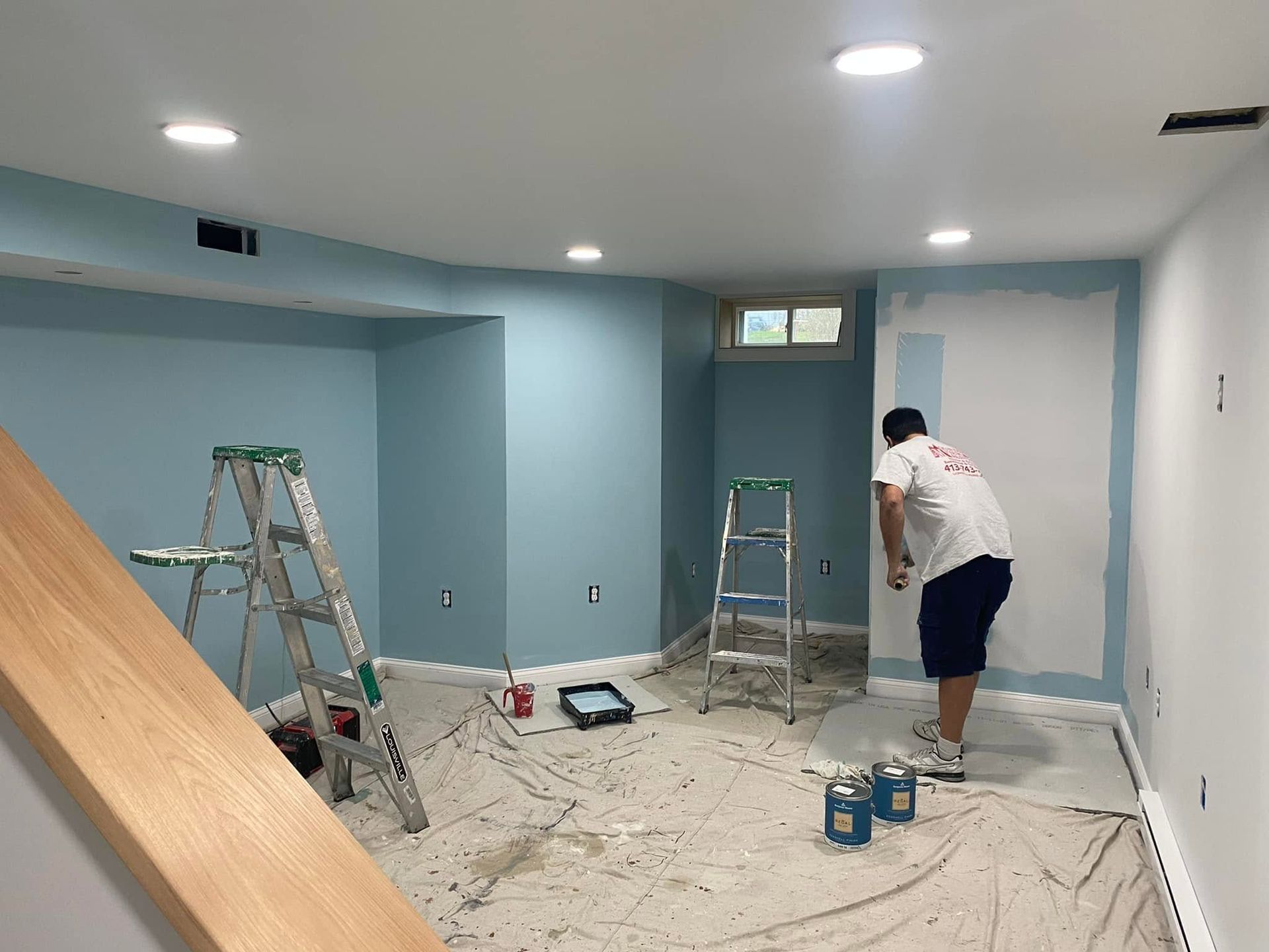Person painting a blue room with ladders, tools, and supplies scattered on the floor.
