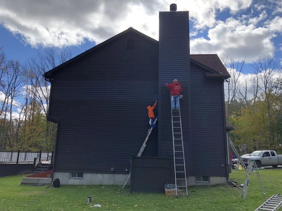 Two workers on ladders repairing a tall chimney on a dark house under cloudy skies.