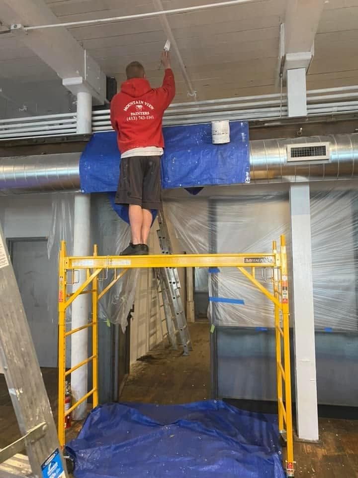 Worker in red shirt standing on yellow scaffolding, installing overhead ceiling panels in a construction area.
