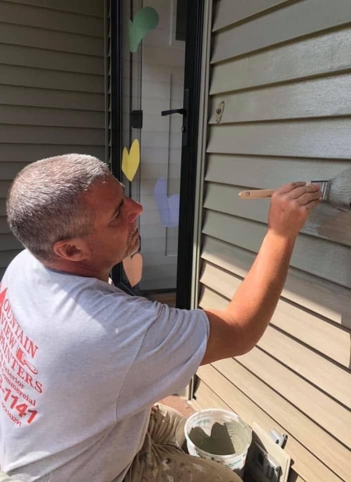 Person painting the exterior siding of a house with a brush, using a bucket of paint nearby.