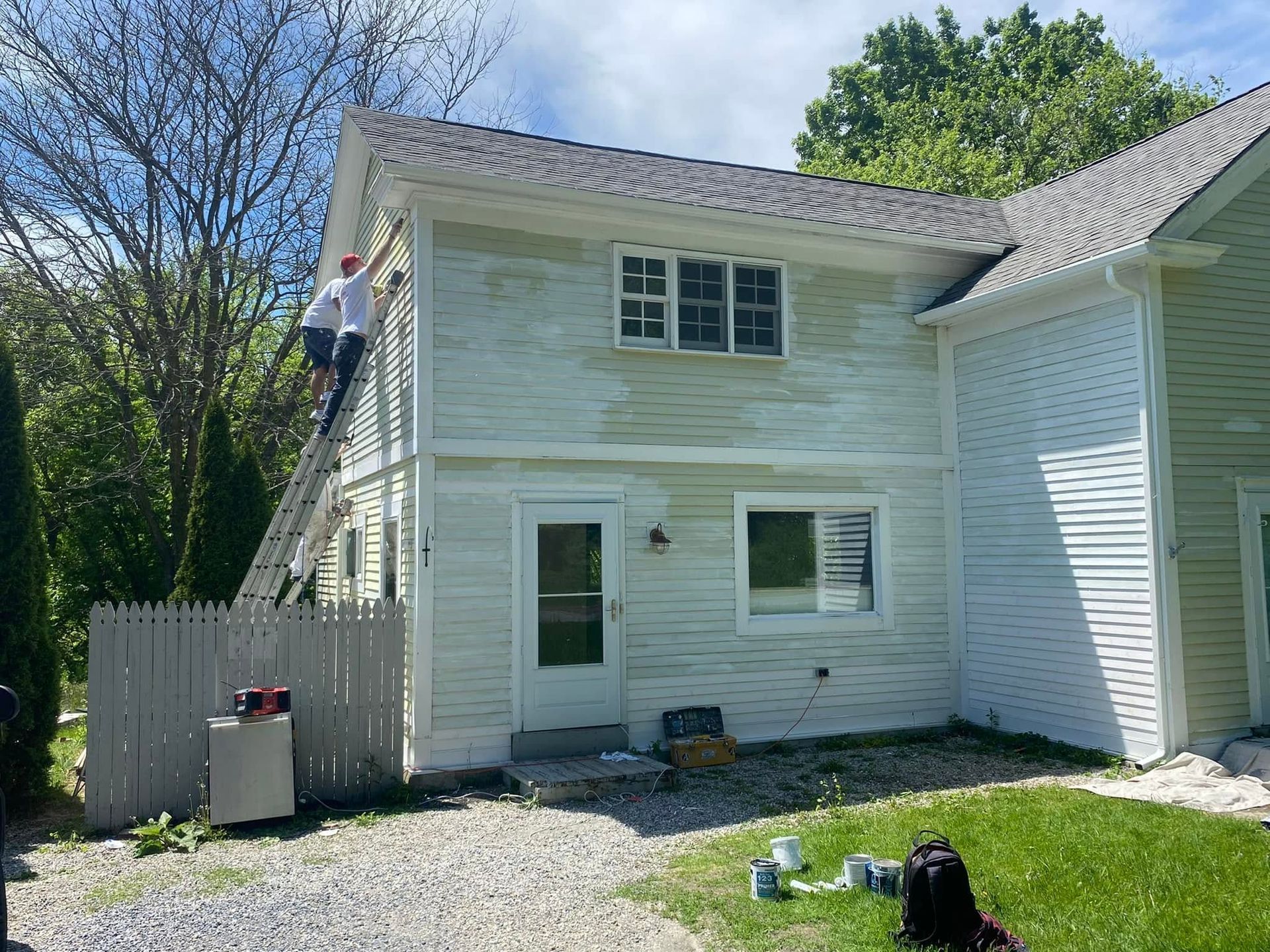 Person on a ladder painting the side of a white house in a sunny yard