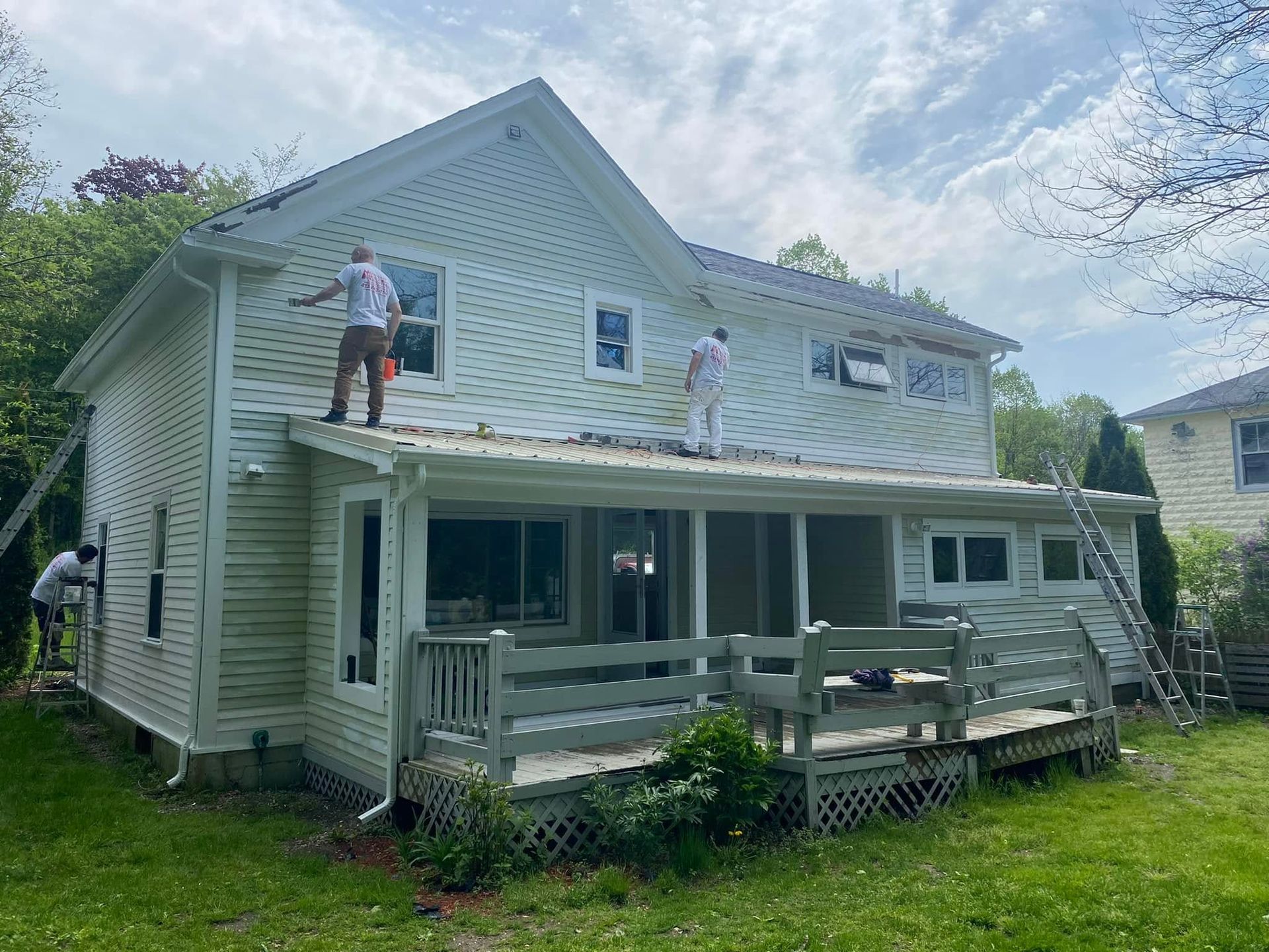 Two people painting a white two-story house with a front porch and ladder under a cloudy sky