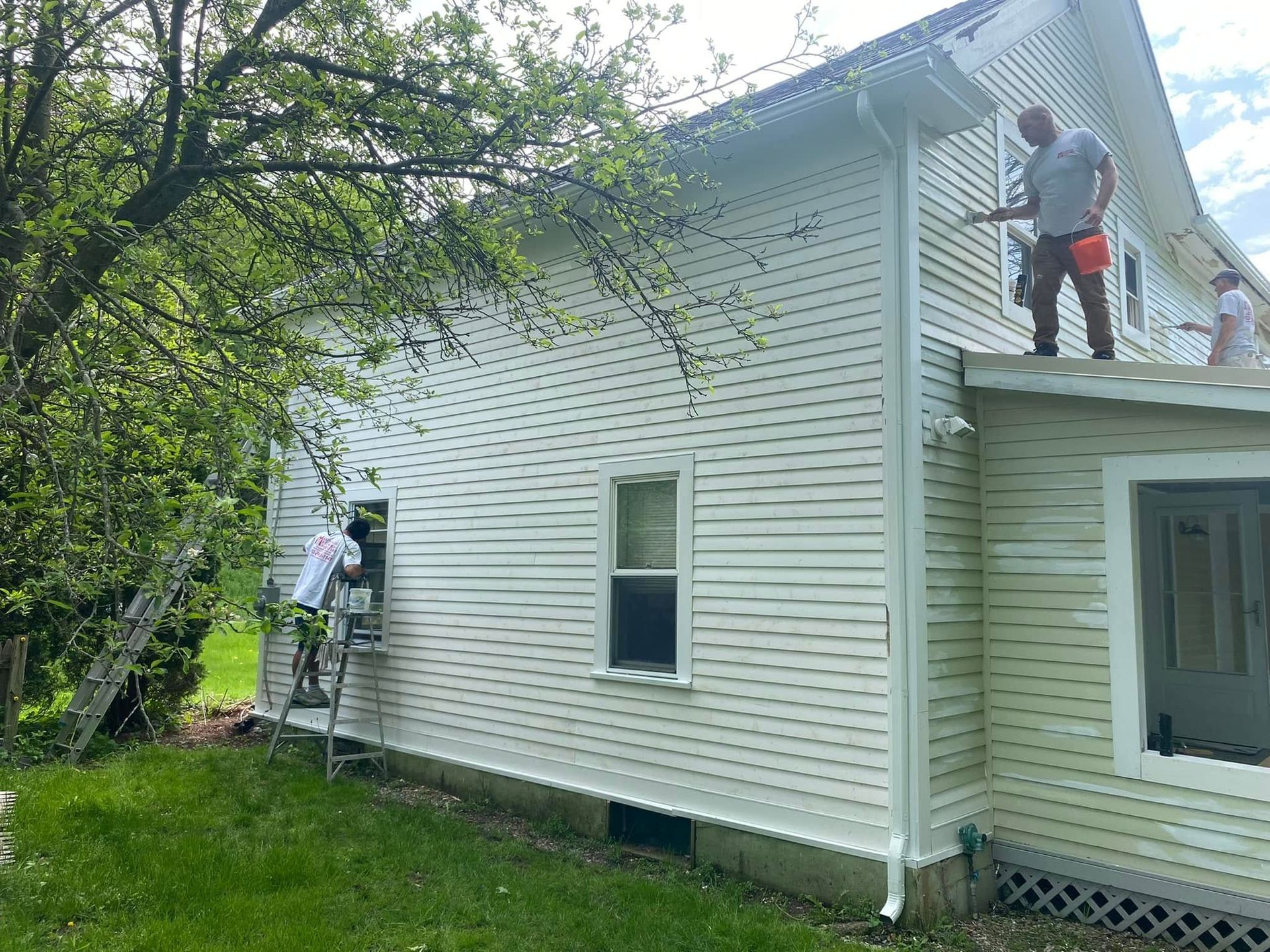 Two workers on ladders repairing the white siding and roof of a house.
