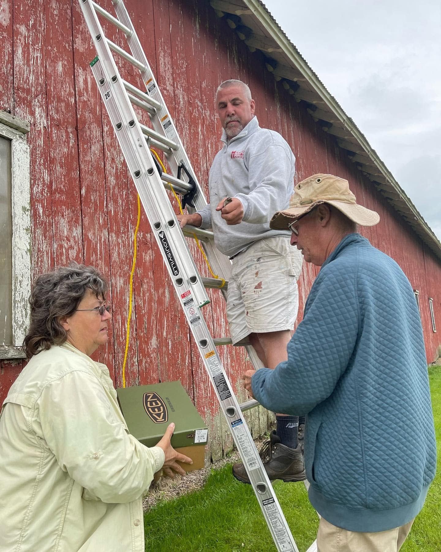 Three people by a red barn, one on a ladder painting while two others assist below.