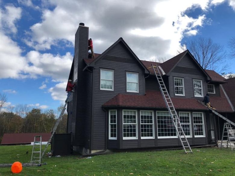 Two ladders lean against a dark house with workers on the roof under a cloudy sky