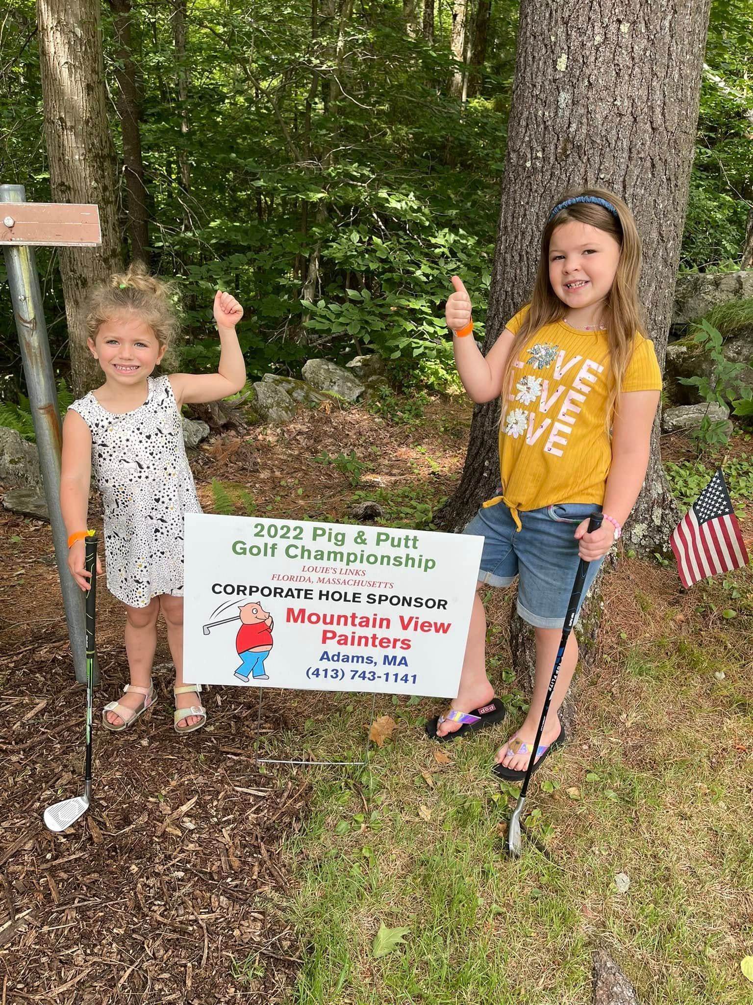 Two children holding a summer camp sign between trees, both giving thumbs up.
