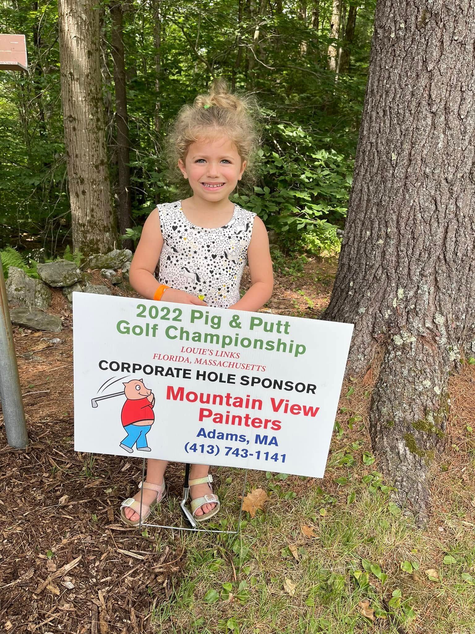 Girl holding a sign for the 2023 Golf & Putt Championship at Corporate Woods Park, outdoors by trees