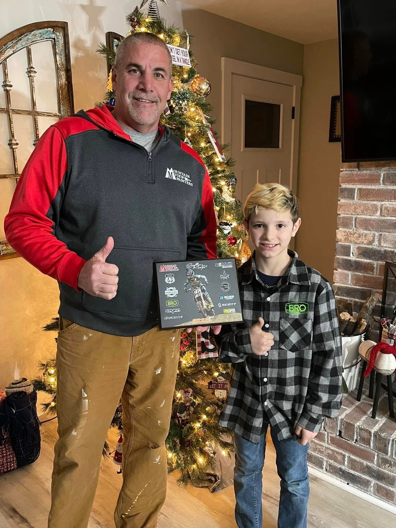 Man and child smiling indoors beside a lit Christmas tree, holding a small chalkboard sign and giving thumbs up