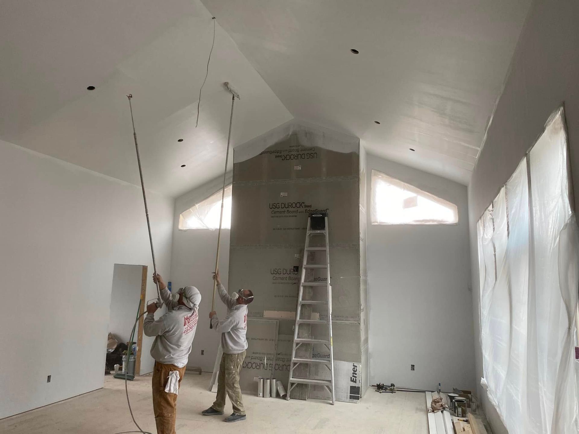 Two workers installing ceiling fixtures in a bright room with a ladder and plastic-covered wall.