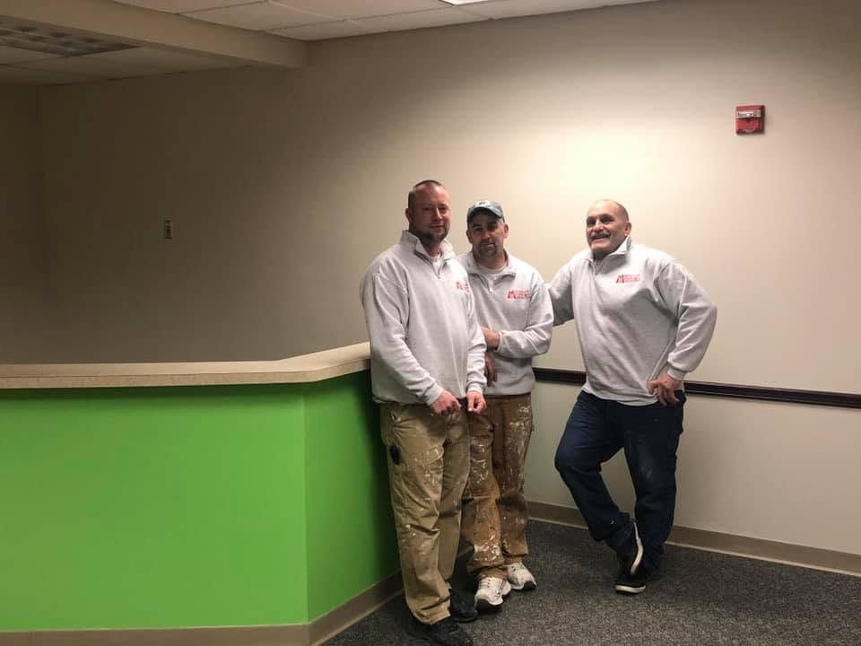 Three people standing by a green reception counter in a beige hallway, smiling and leaning casually