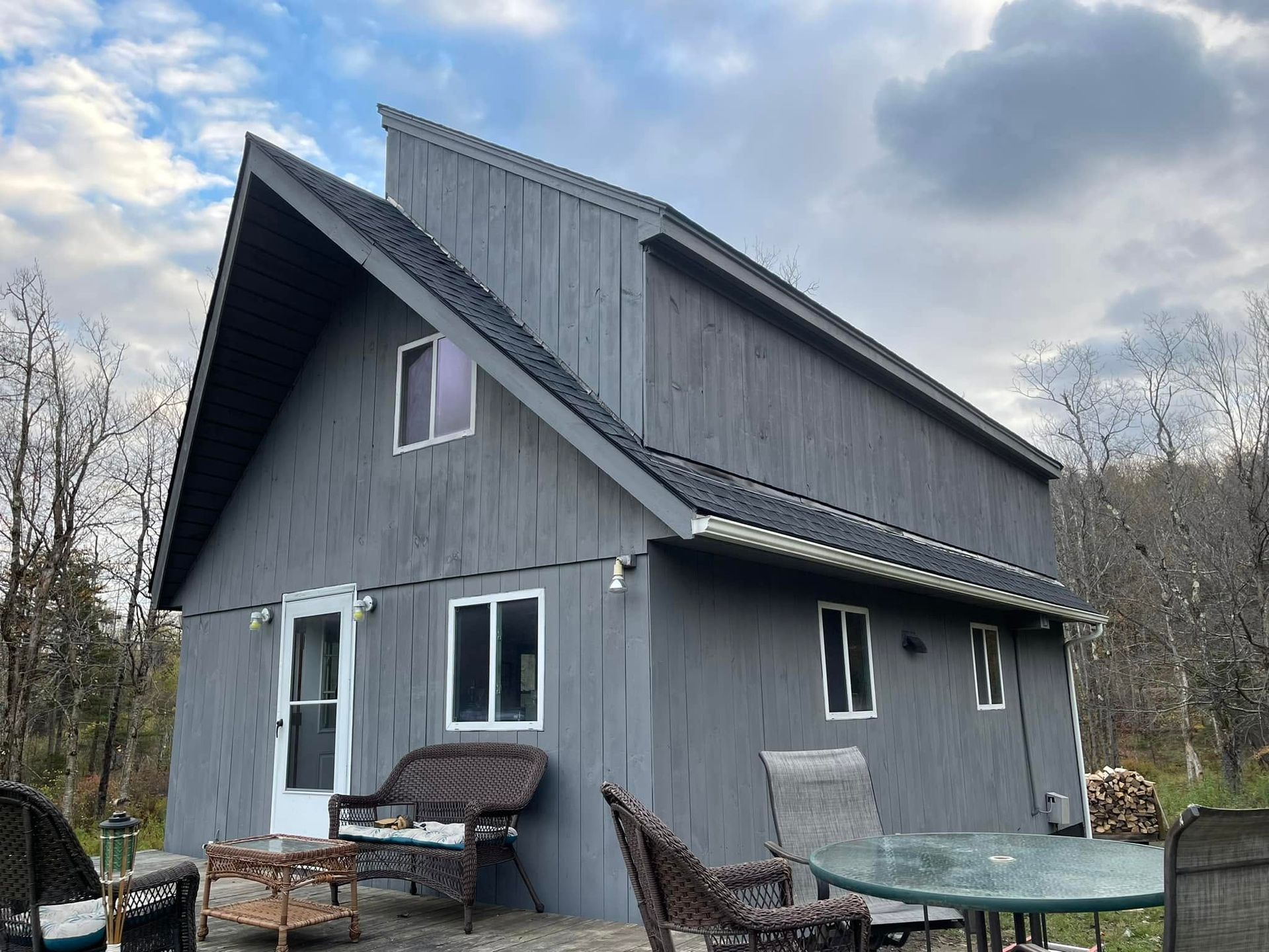 Gray A-frame cabin with a patio set under a cloudy sky in a wooded area