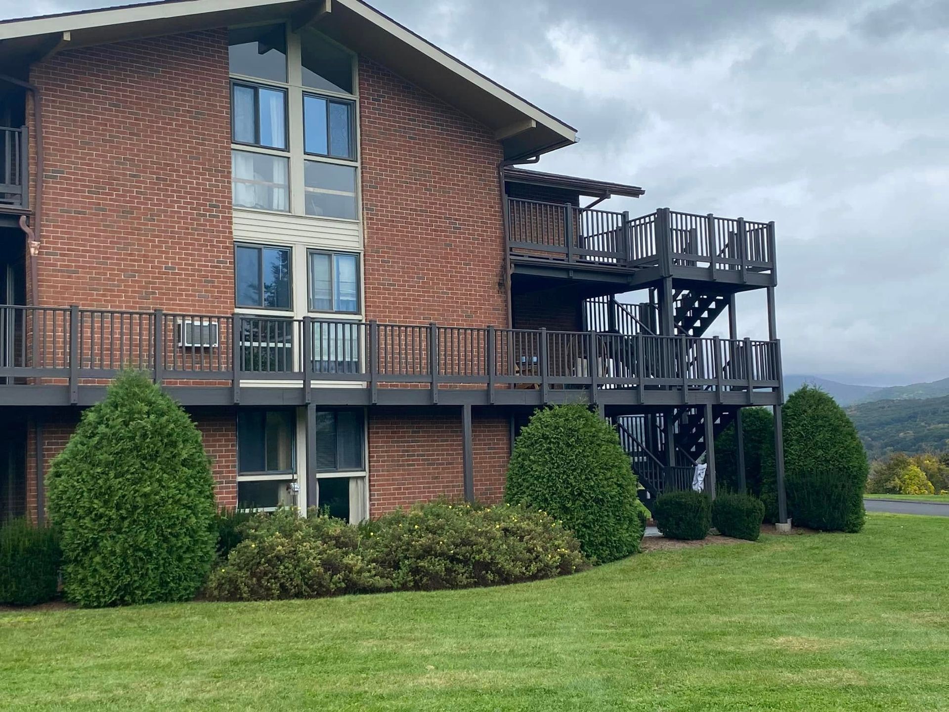 Brick house with wooden decks and stairs, surrounded by a green lawn under cloudy skies