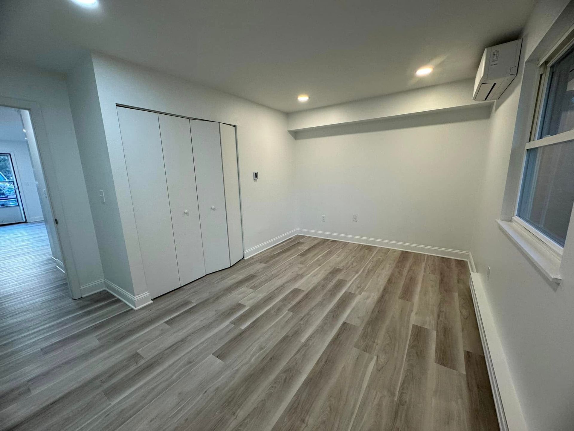 Empty modern room with gray wood floors, white walls, closet doors, and a window.