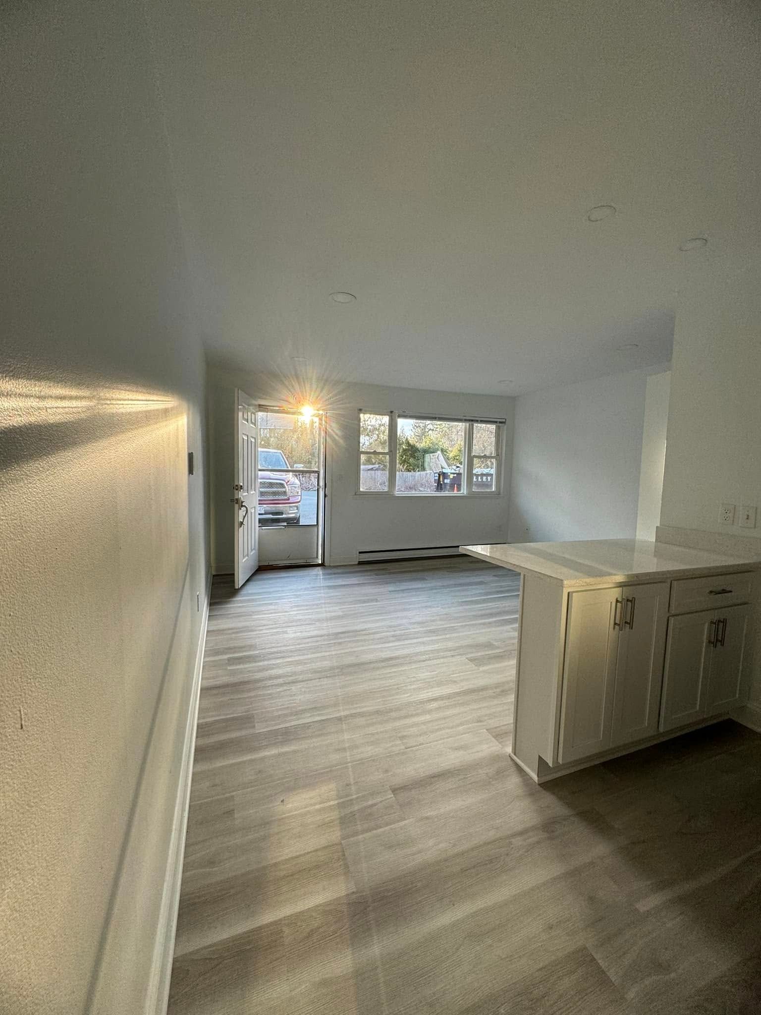 Empty white living room with gray wood floors, kitchen island, and large window at the far end.