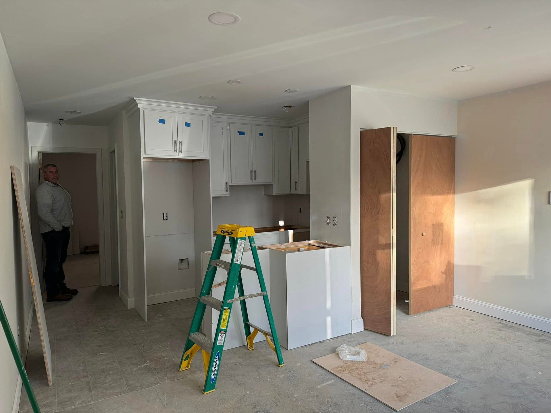 Small room under renovation with cabinets, a ladder, and open closet doors; a person stands in the doorway.