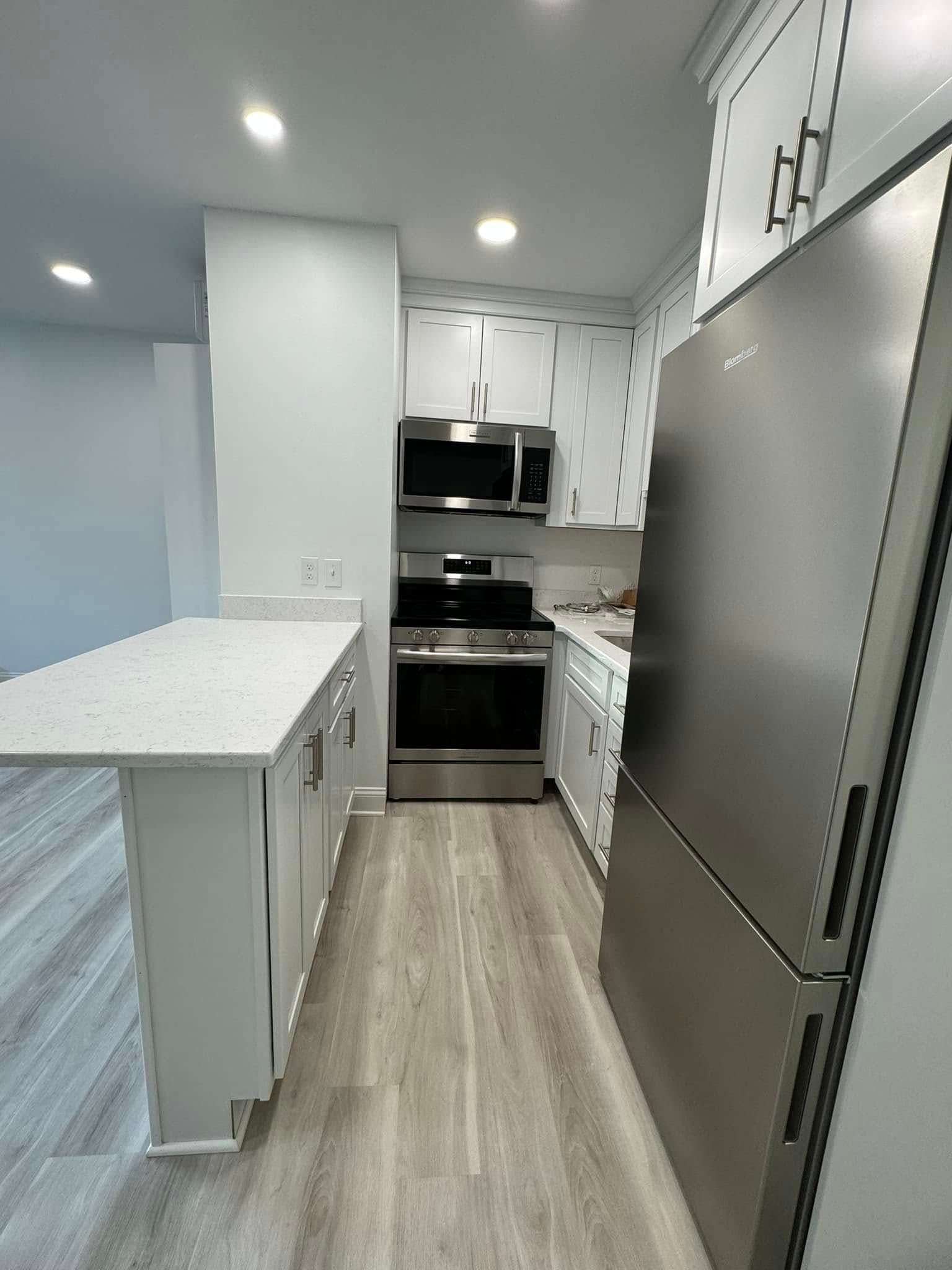 Narrow modern kitchen with white cabinets, stainless steel refrigerator, and wood-look flooring.