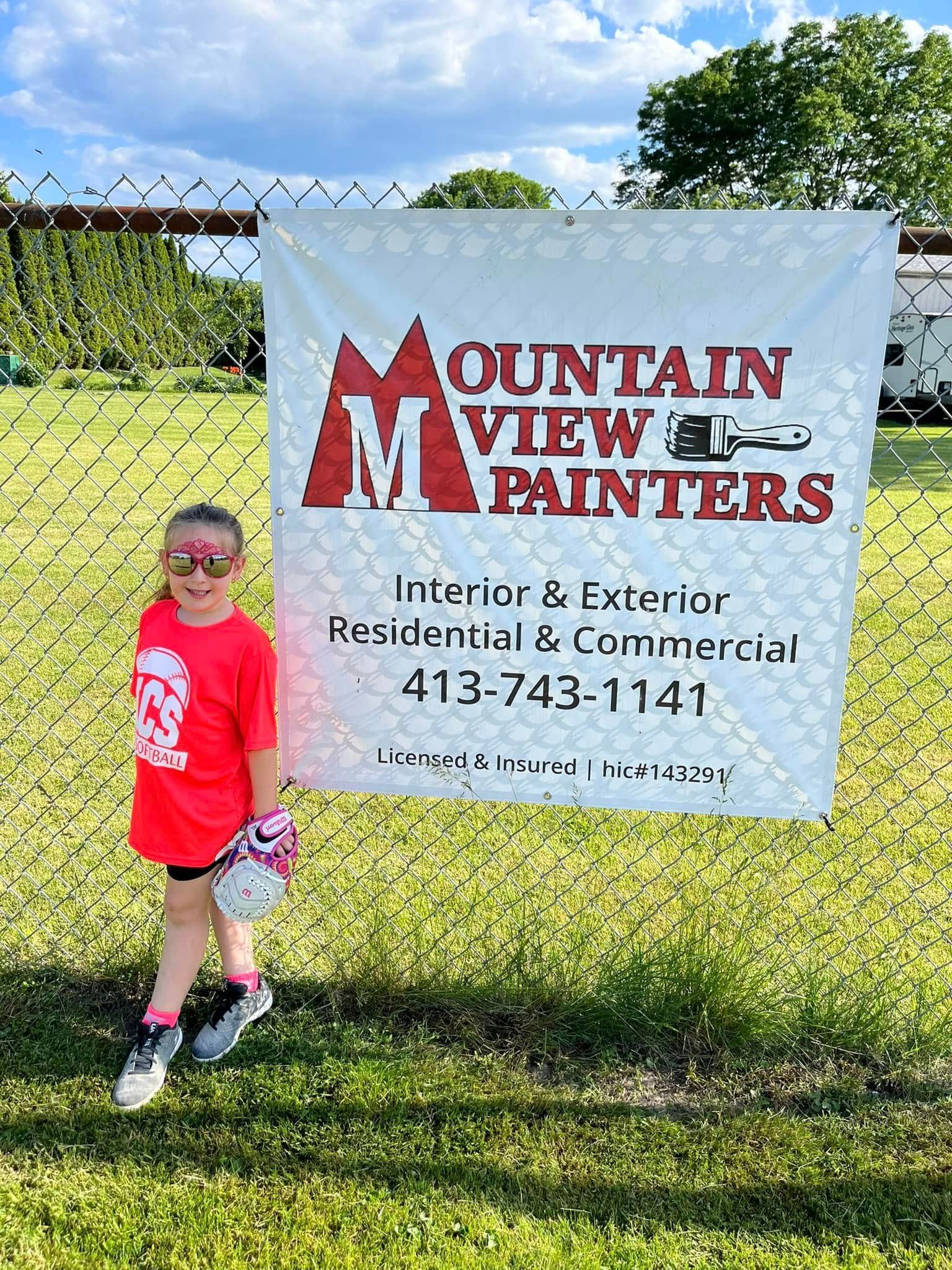 Child in red shirt standing beside a Mountain View Painters banner on a grassy field.