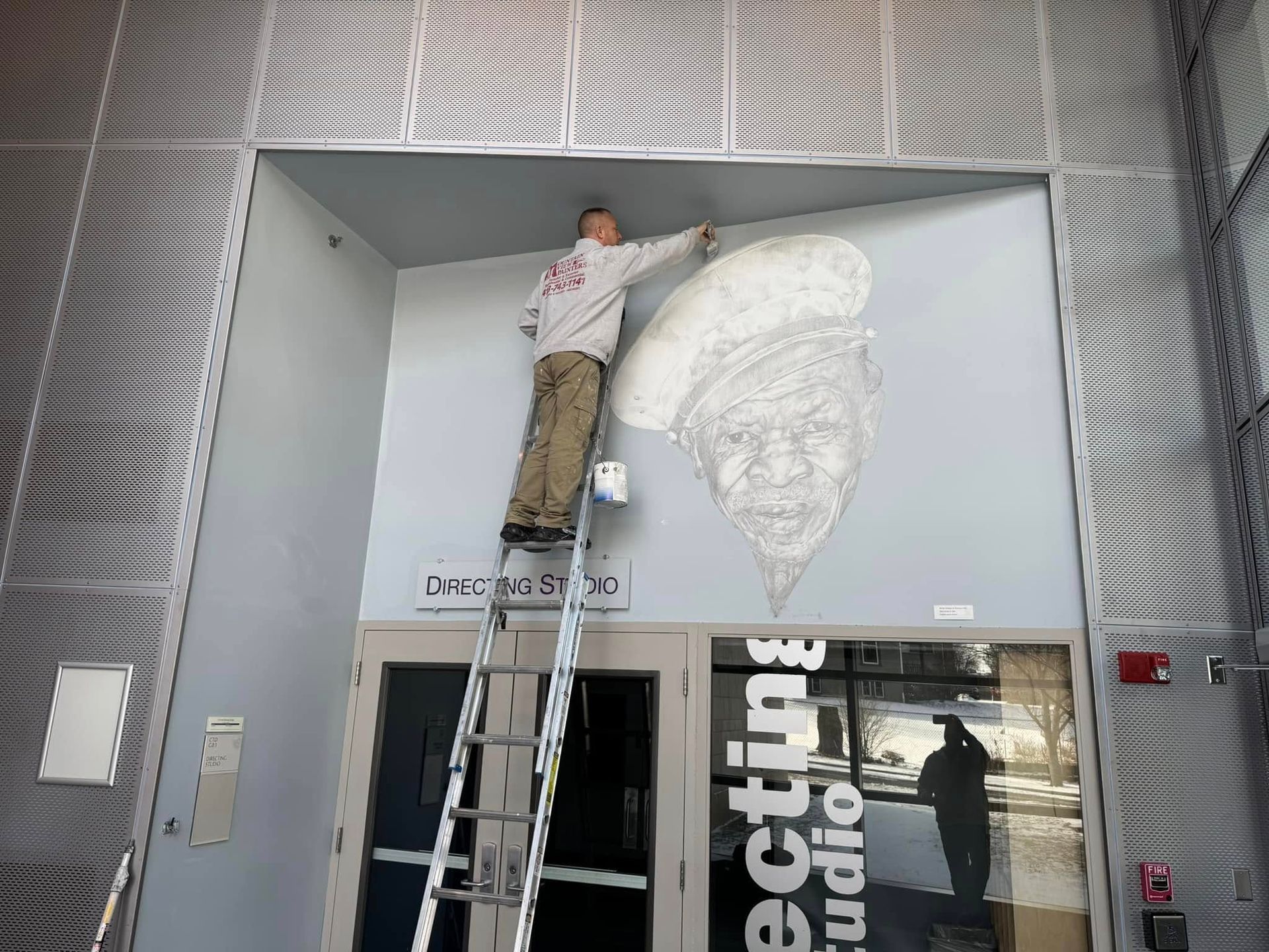 Person on a ladder painting a large white wall mural inside a modern building.