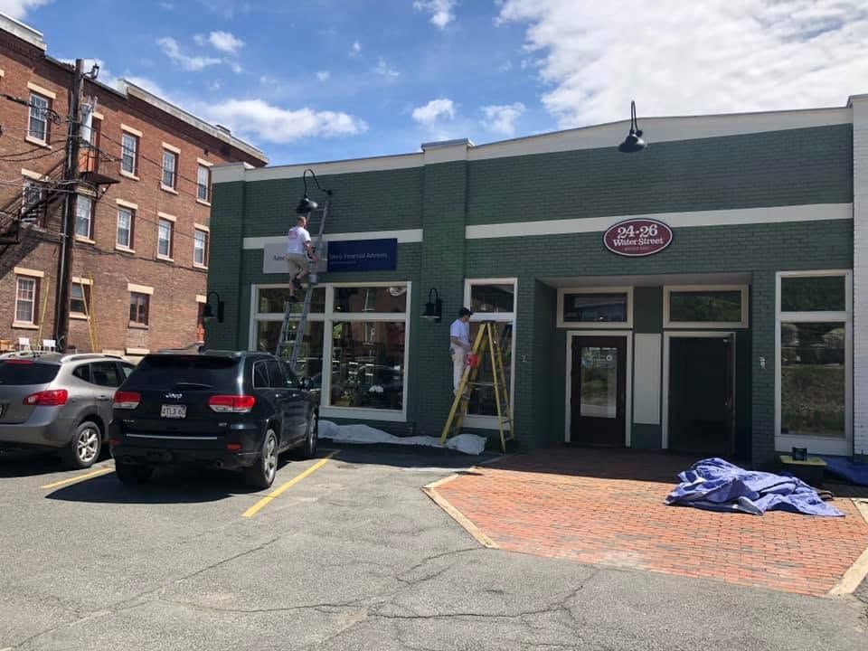 Storefronts on a sunny street, with a black SUV parked outside and a ladder by the entrance.