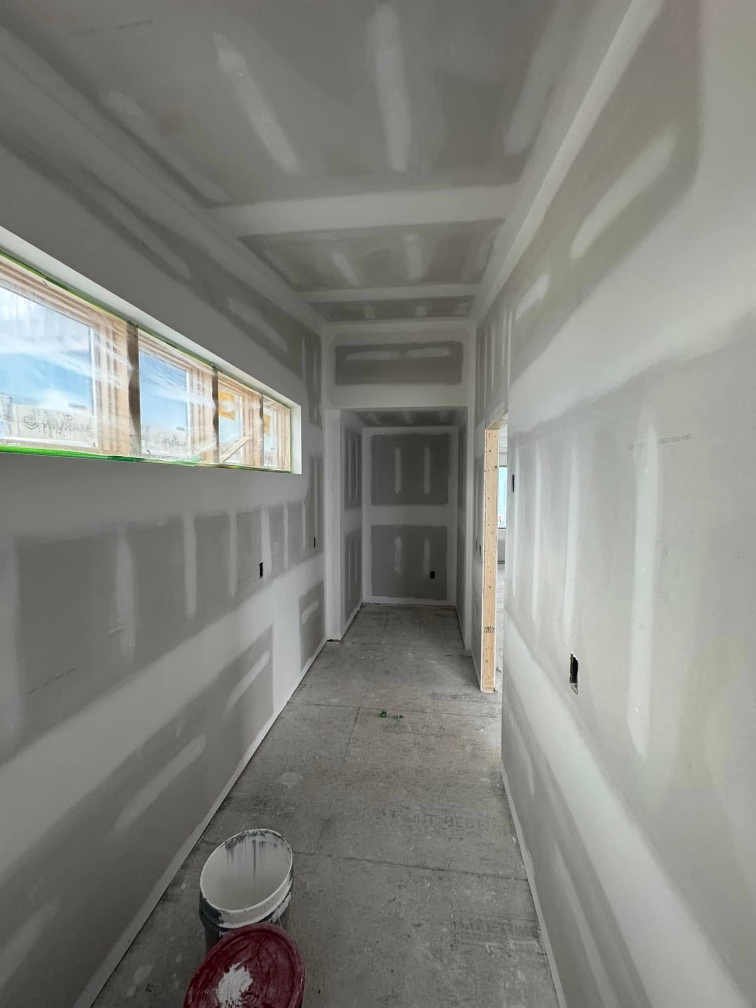 Narrow unfinished hallway with drywall, windows on the left, and a paint bucket on the floor.