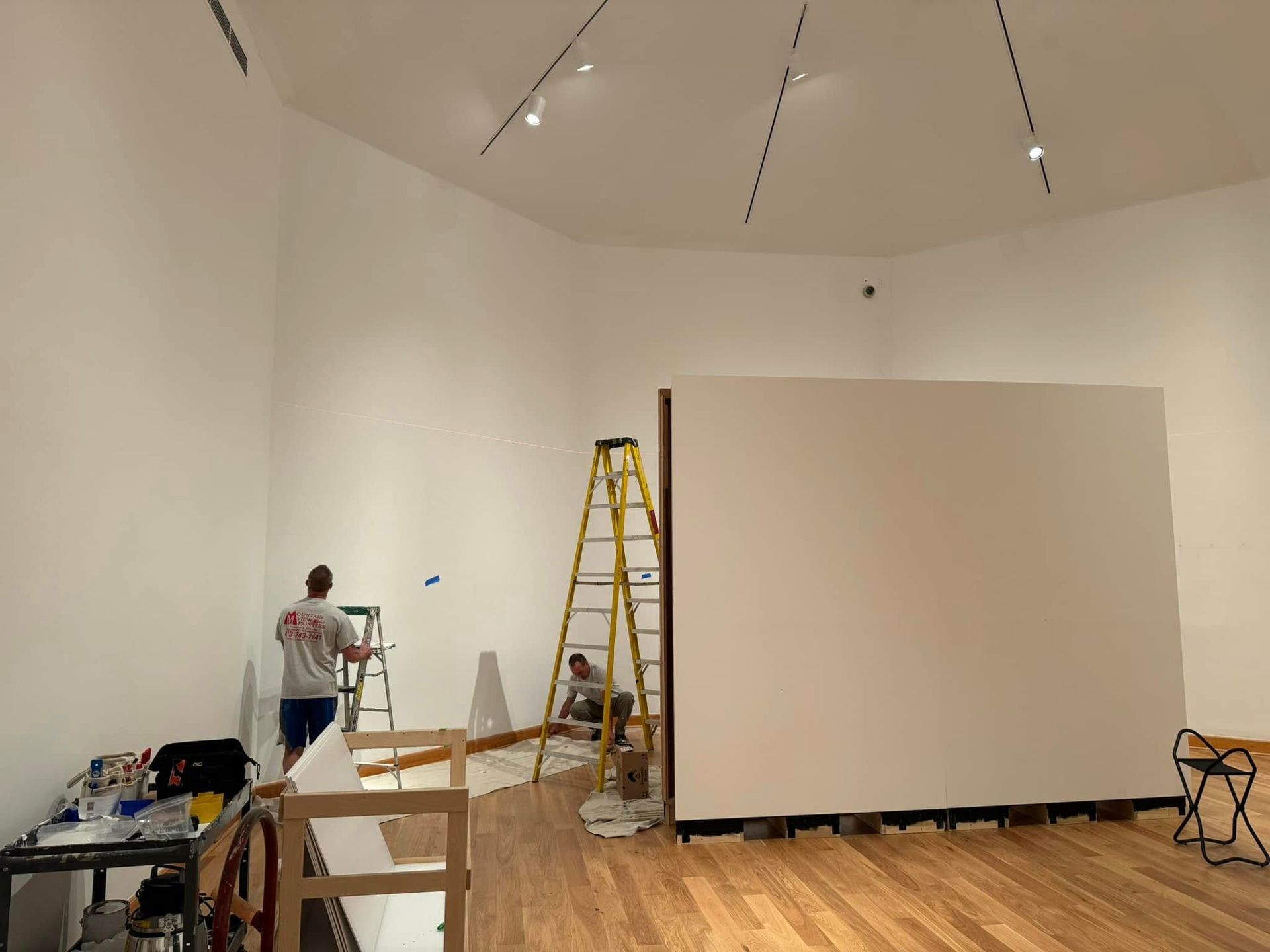 Gallery room with white walls, a large partition, ladder, and two people painting or installing fixtures