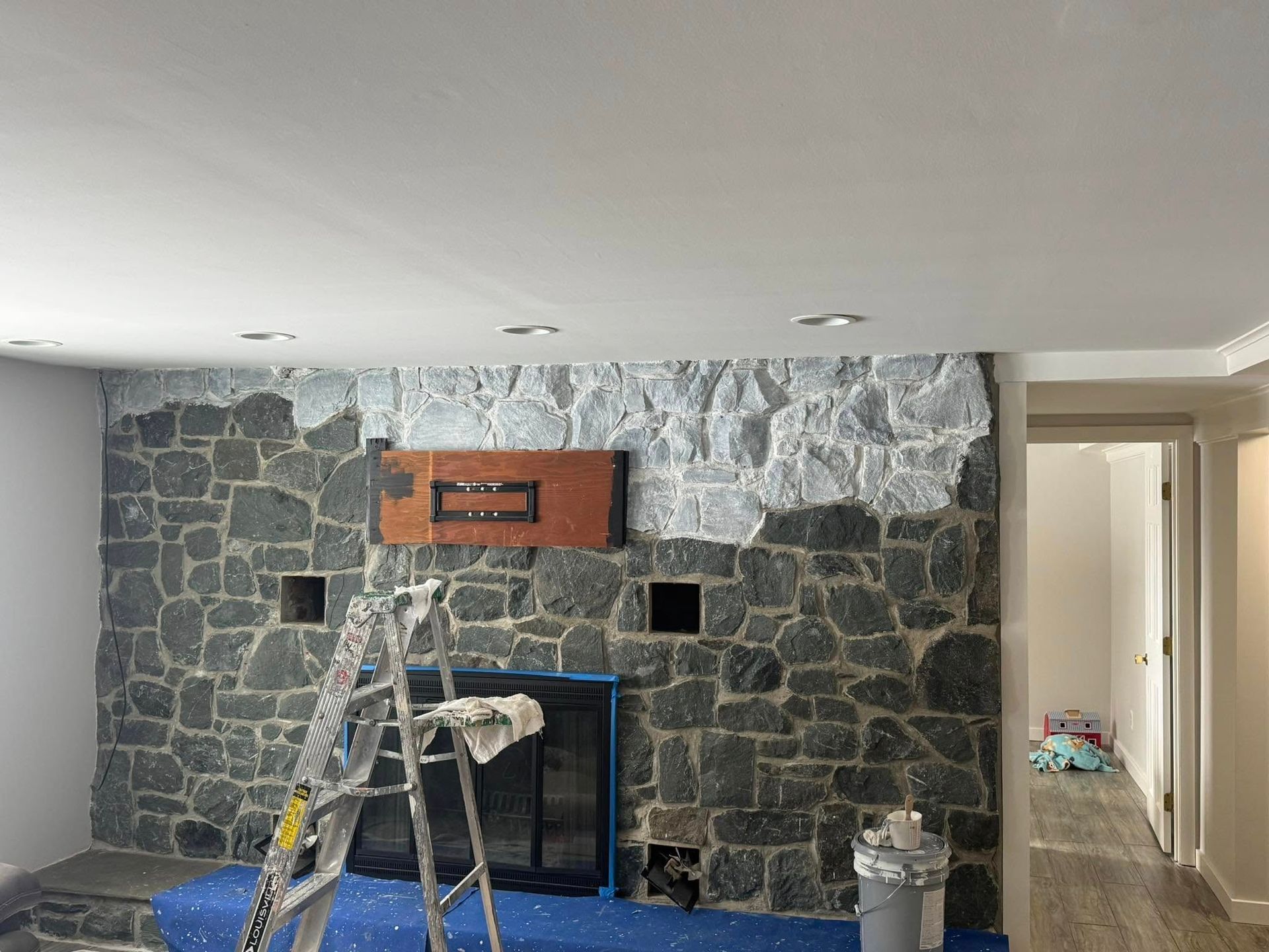Stone fireplace under renovation with a ladder, paint supplies, and hallway visible on the right.