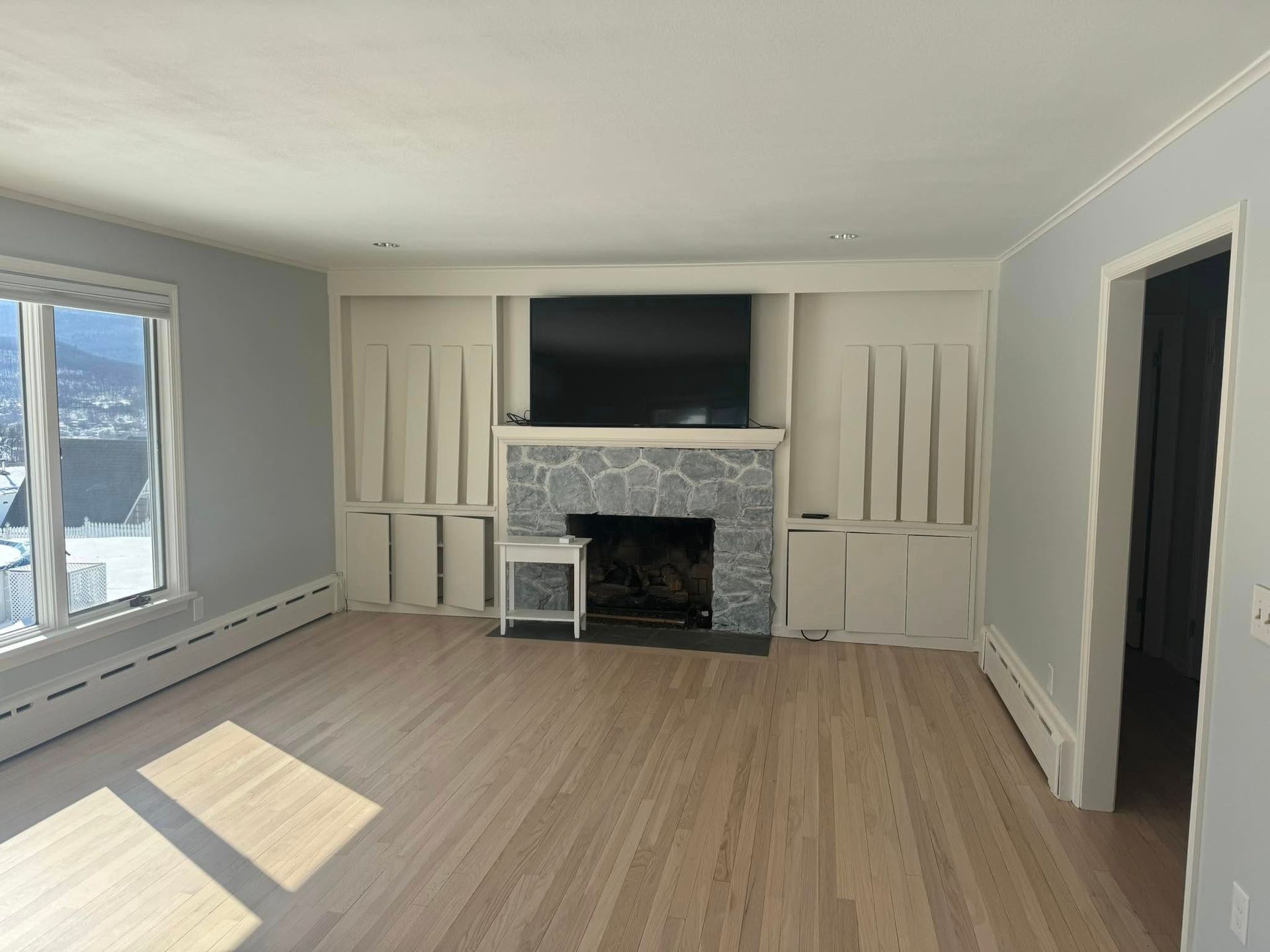 Empty living room with stone fireplace, built-in cabinets, large windows, and light wood floors.