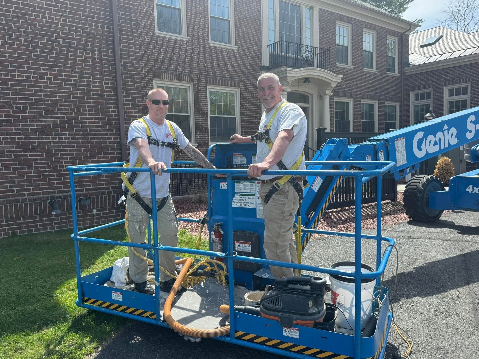 Two workers on a blue Genie lift outside a brick building, with tools and equipment on the platform.
