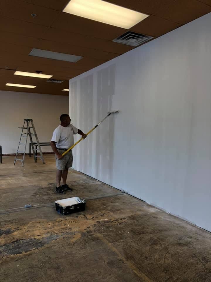 Person painting a large white wall inside a room with a paint roller and ladder nearby