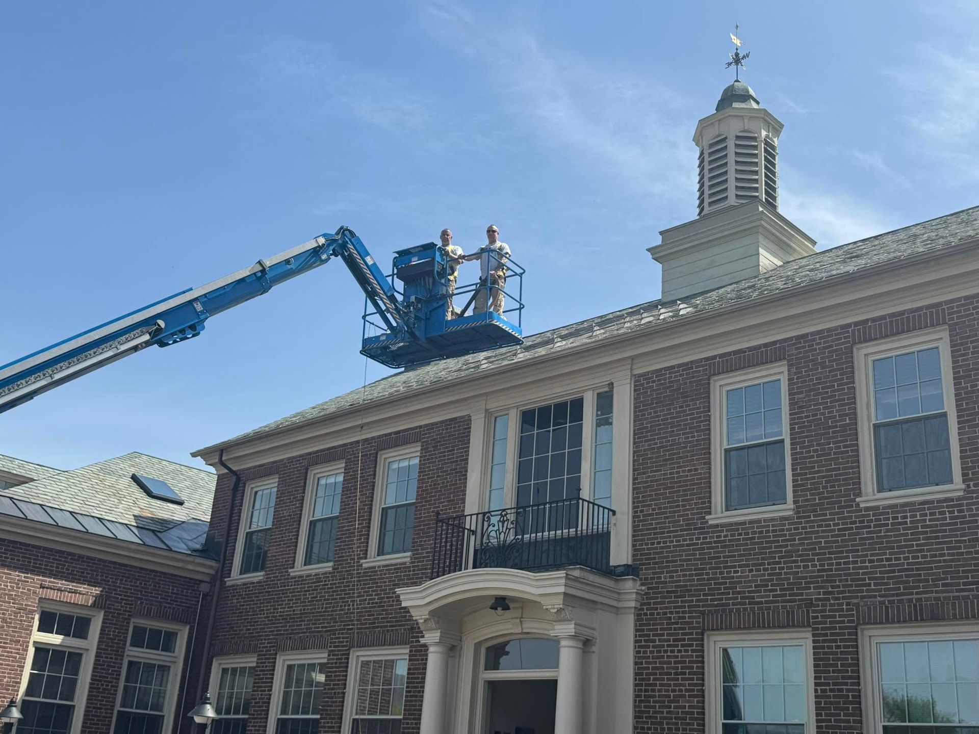 Workers on a blue lift inspect the roof of a brick building with a small cupola.