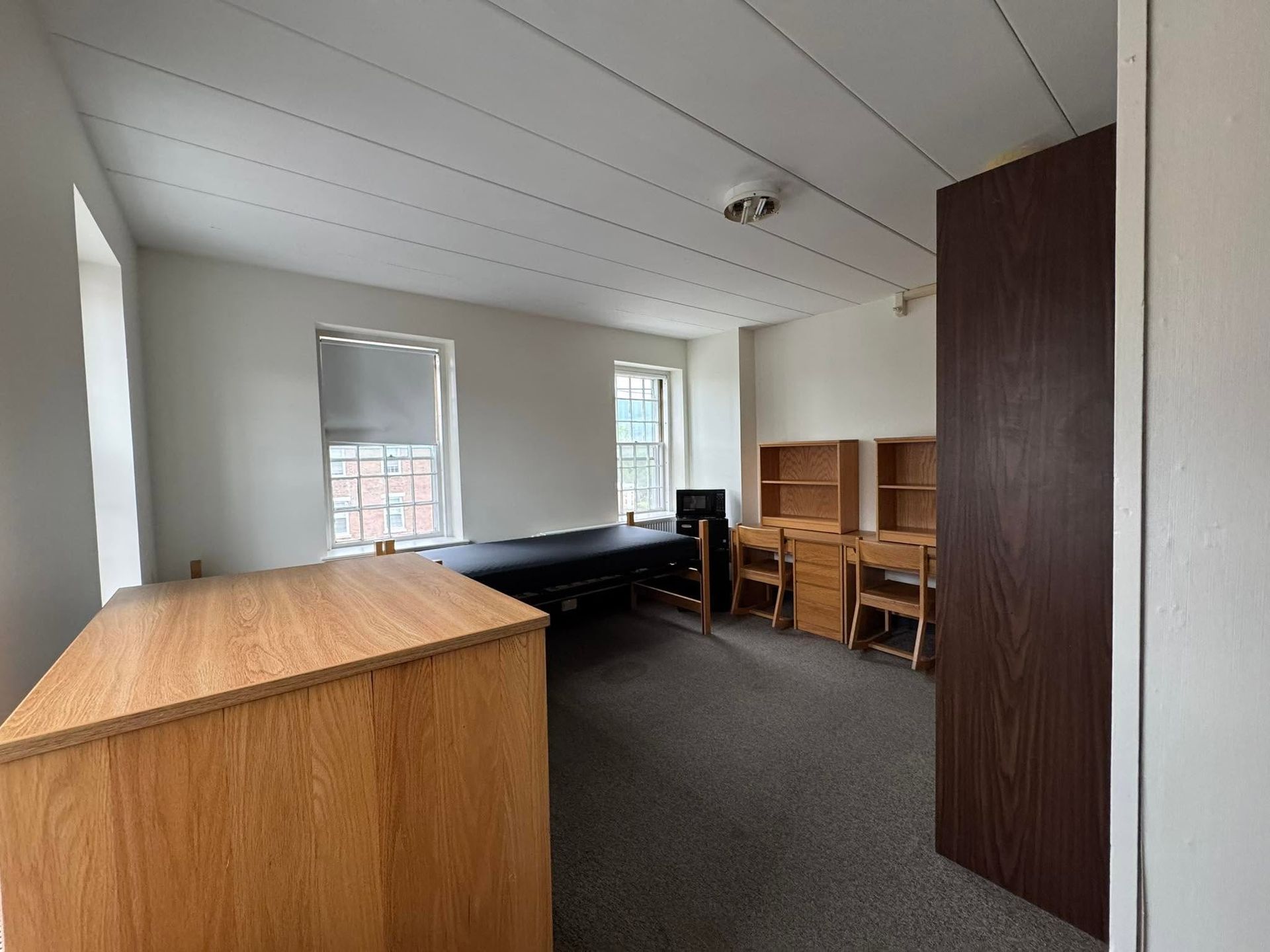 Sparse bedroom with wooden furniture, a bed, desk, chairs, and open dark wardrobe door.