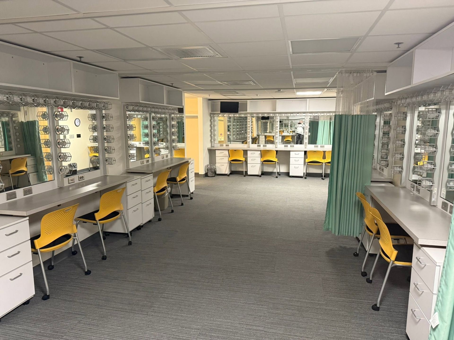 Empty dressing room with mirrored vanities, yellow chairs, and teal curtains under bright lights