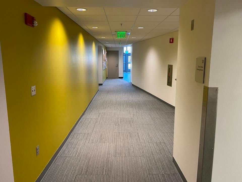Empty office hallway with yellow and white walls, gray carpet, and an exit sign at the end.