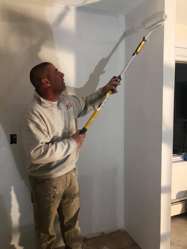 Man painting a white wall with a roller on an extension pole in a room under renovation