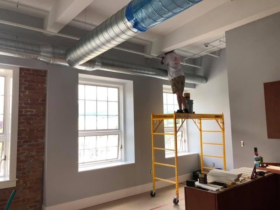 Worker on a yellow scaffold repairing HVAC ducting in a gray room with exposed ceiling beams.