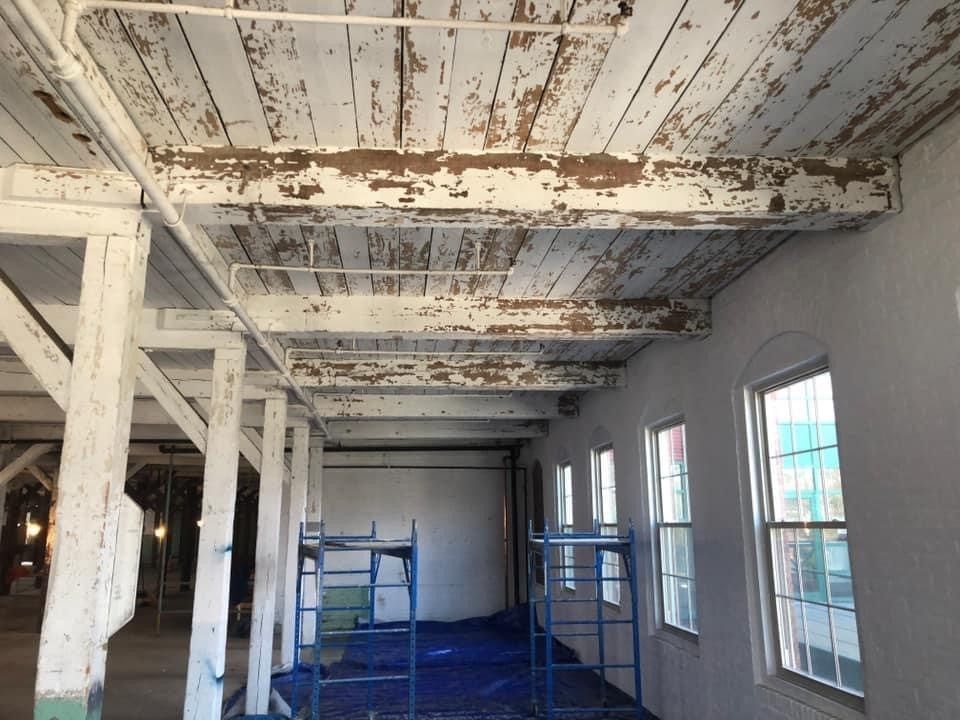 Renovated room with exposed beams, peeling white ceiling, blue tarp floor, and windows along one wall