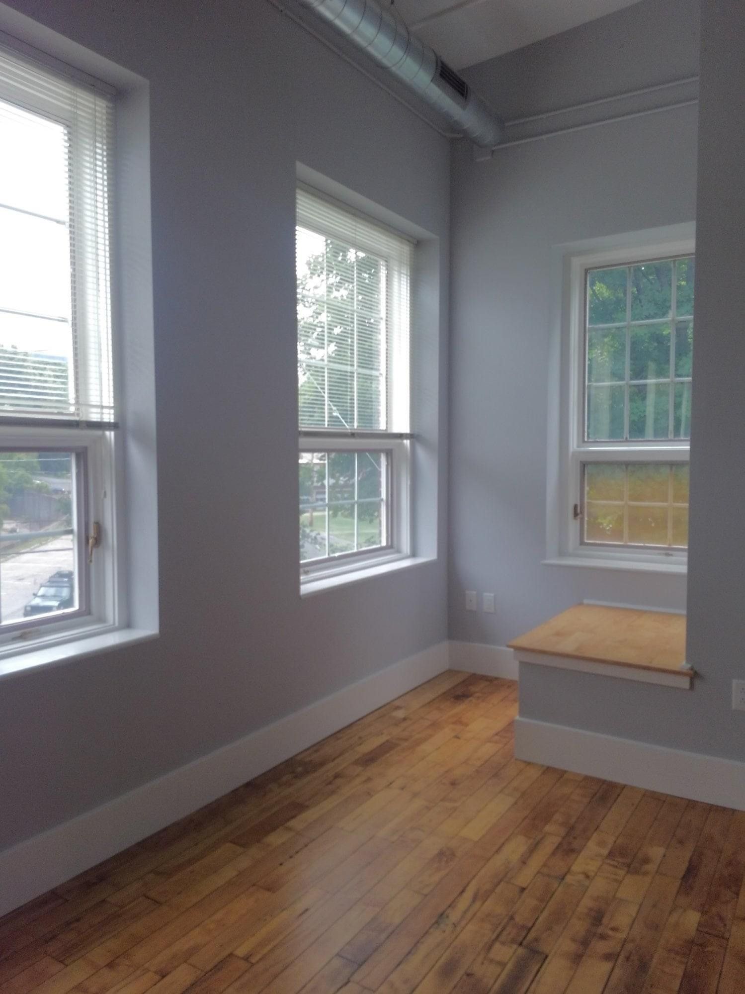 Bright empty sunroom with hardwood floors, white walls, and three large windows, plus a small built-in bench.