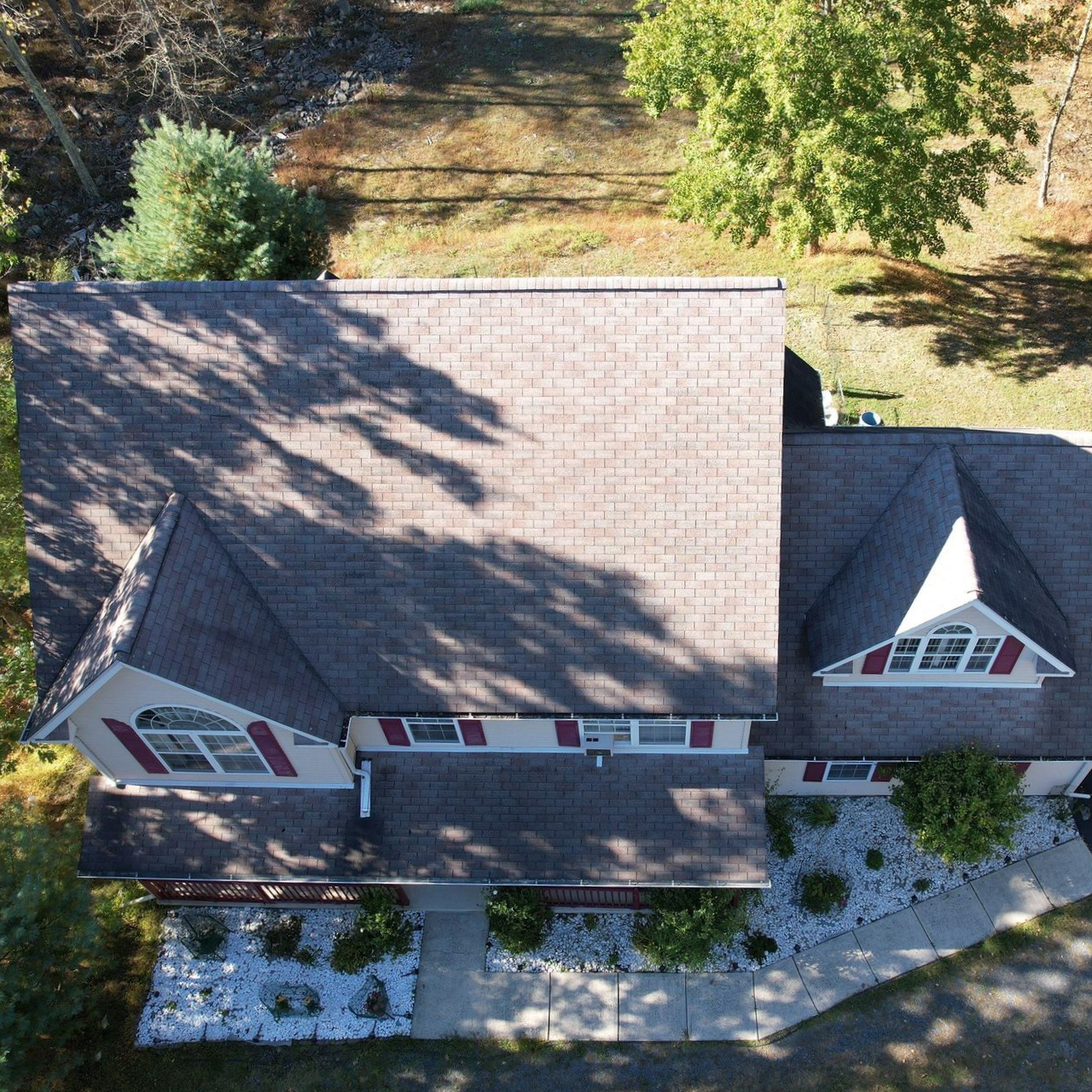 Aerial view of a house under construction with a gray shingle roof and green sheathing. Aerial view of a house with a brown roof, dormer windows, and a landscaped front yard.