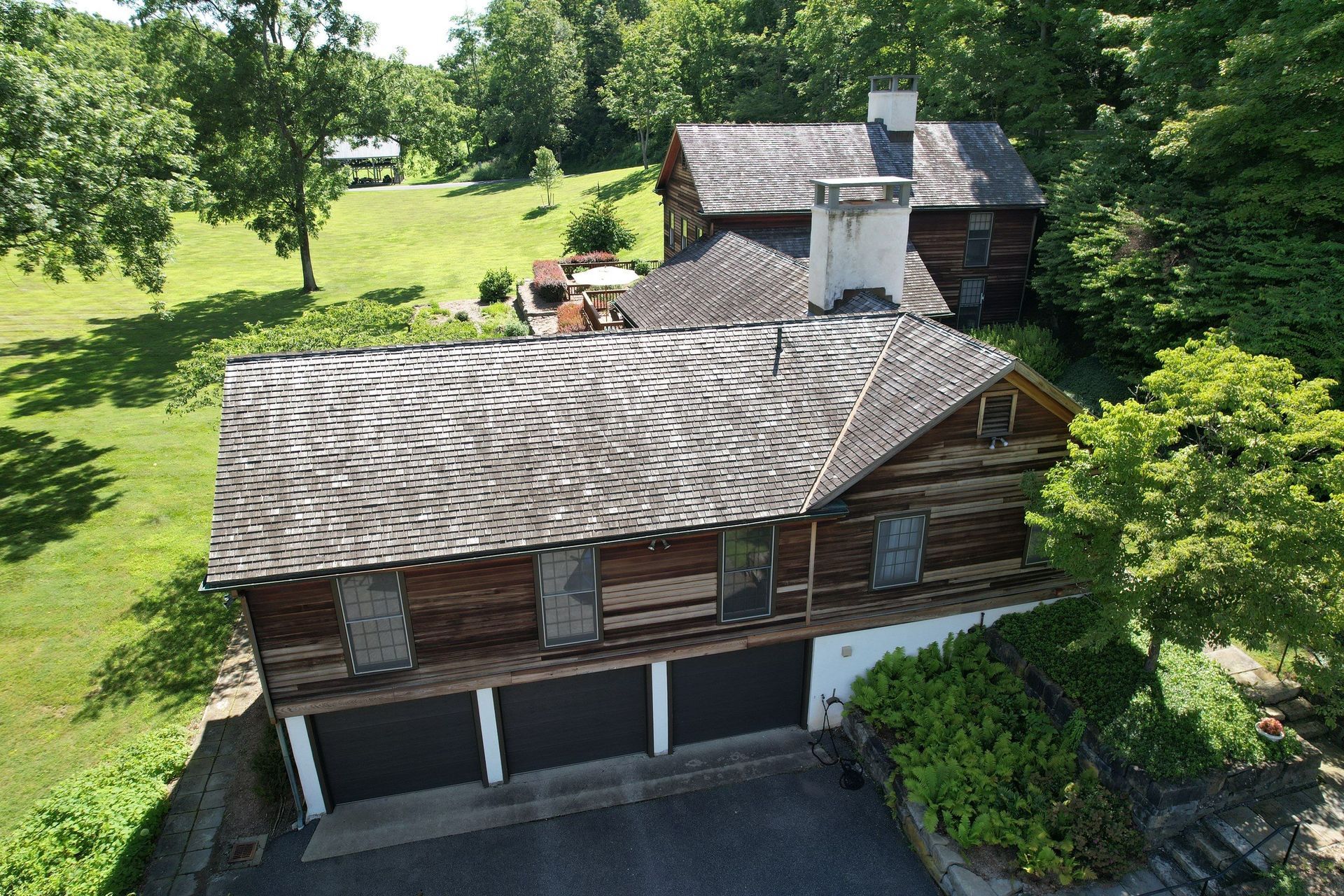 Log cabin house with a garage and wooden roof, nestled in a green, wooded area.