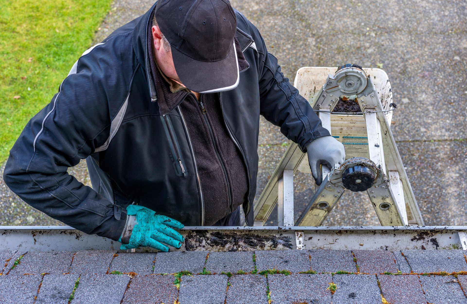 Roof worker cleaning debris from a gutter with a ladder and tools on a house roof