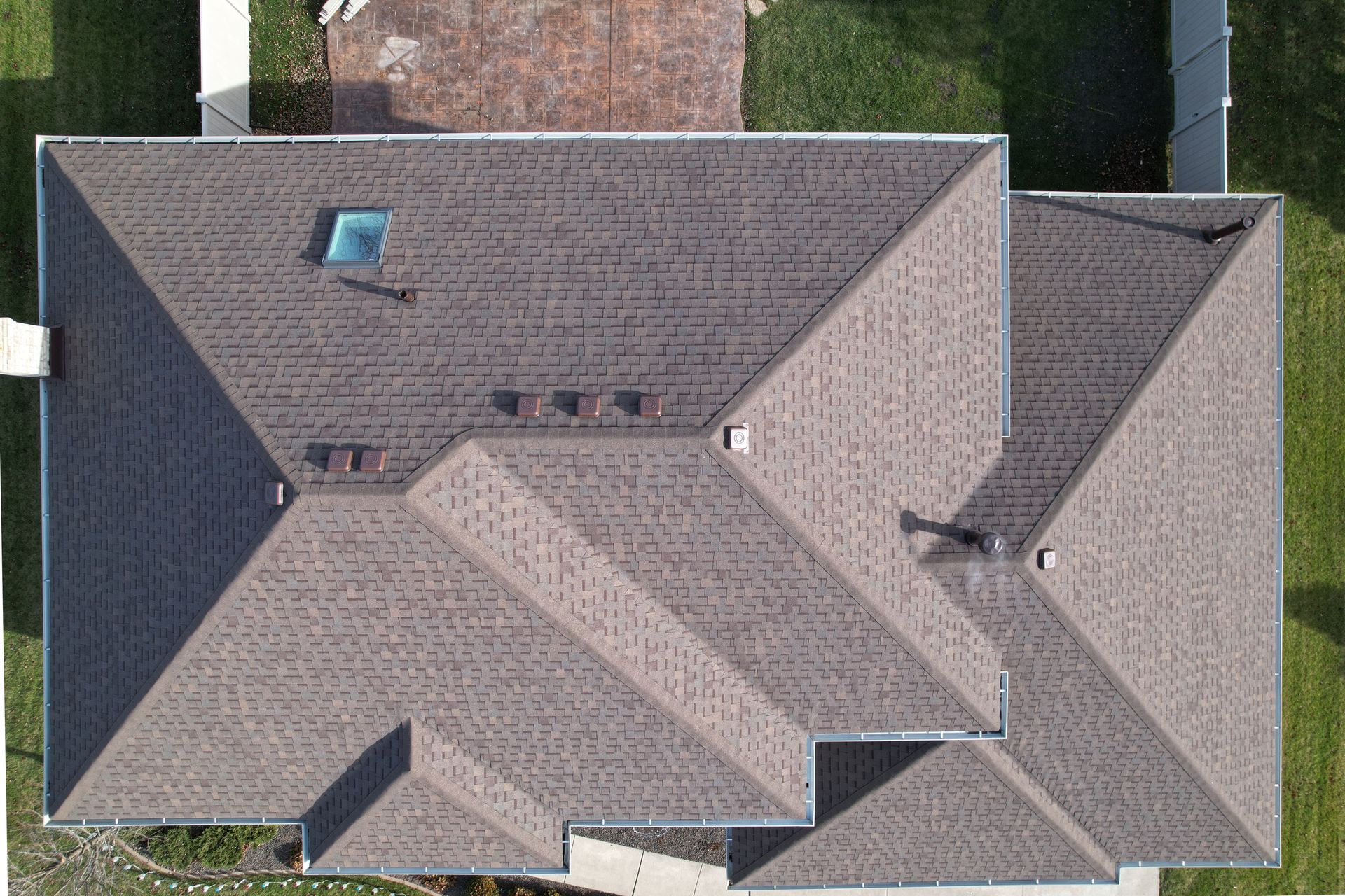 Aerial view of a house roof with gray shingles, skylight, and chimneys surrounded by trees and lawn.