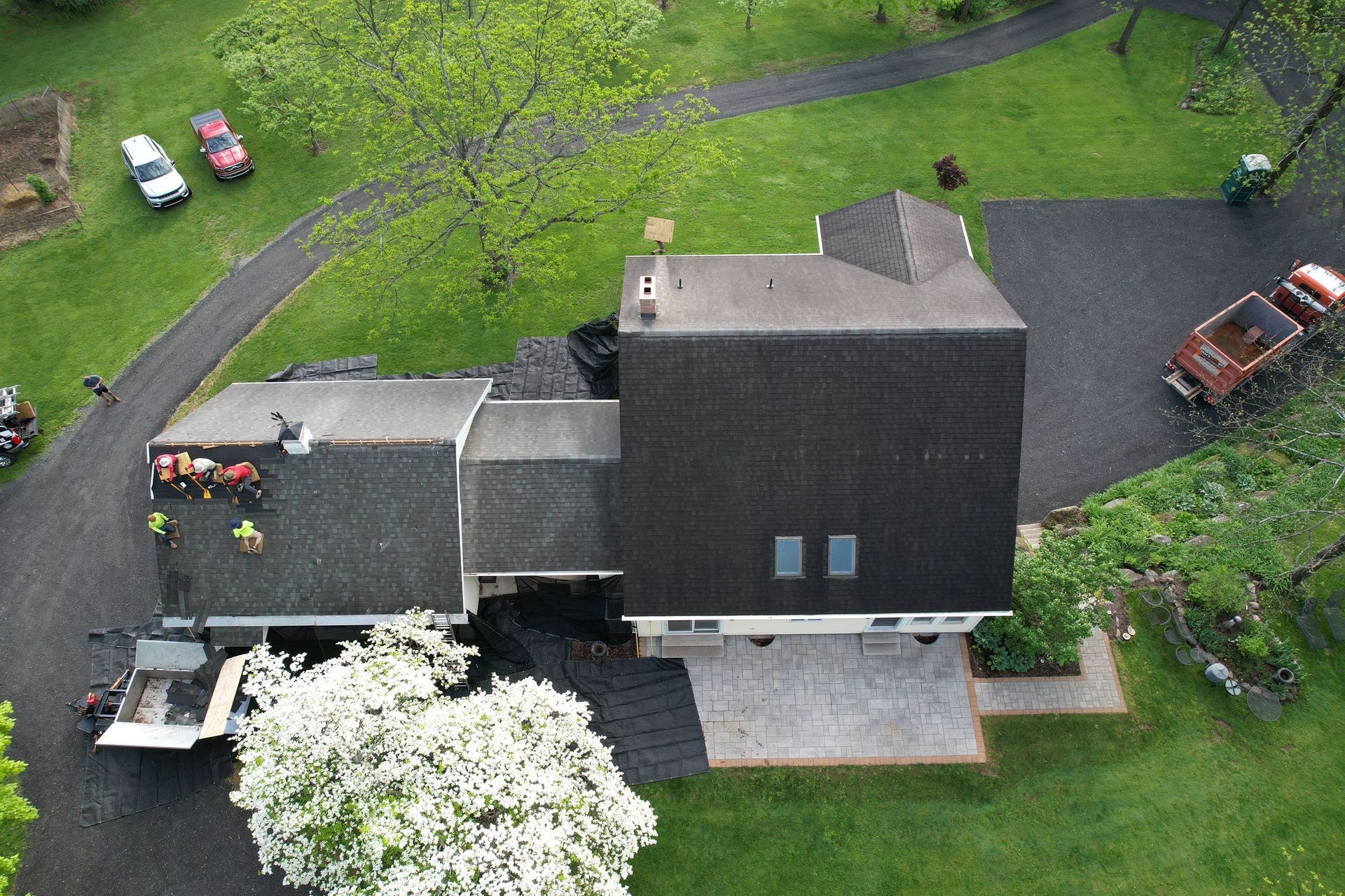 Overhead view of a house; workers on roof. Cars and a truck are in the driveway. Lush green yard surrounds.